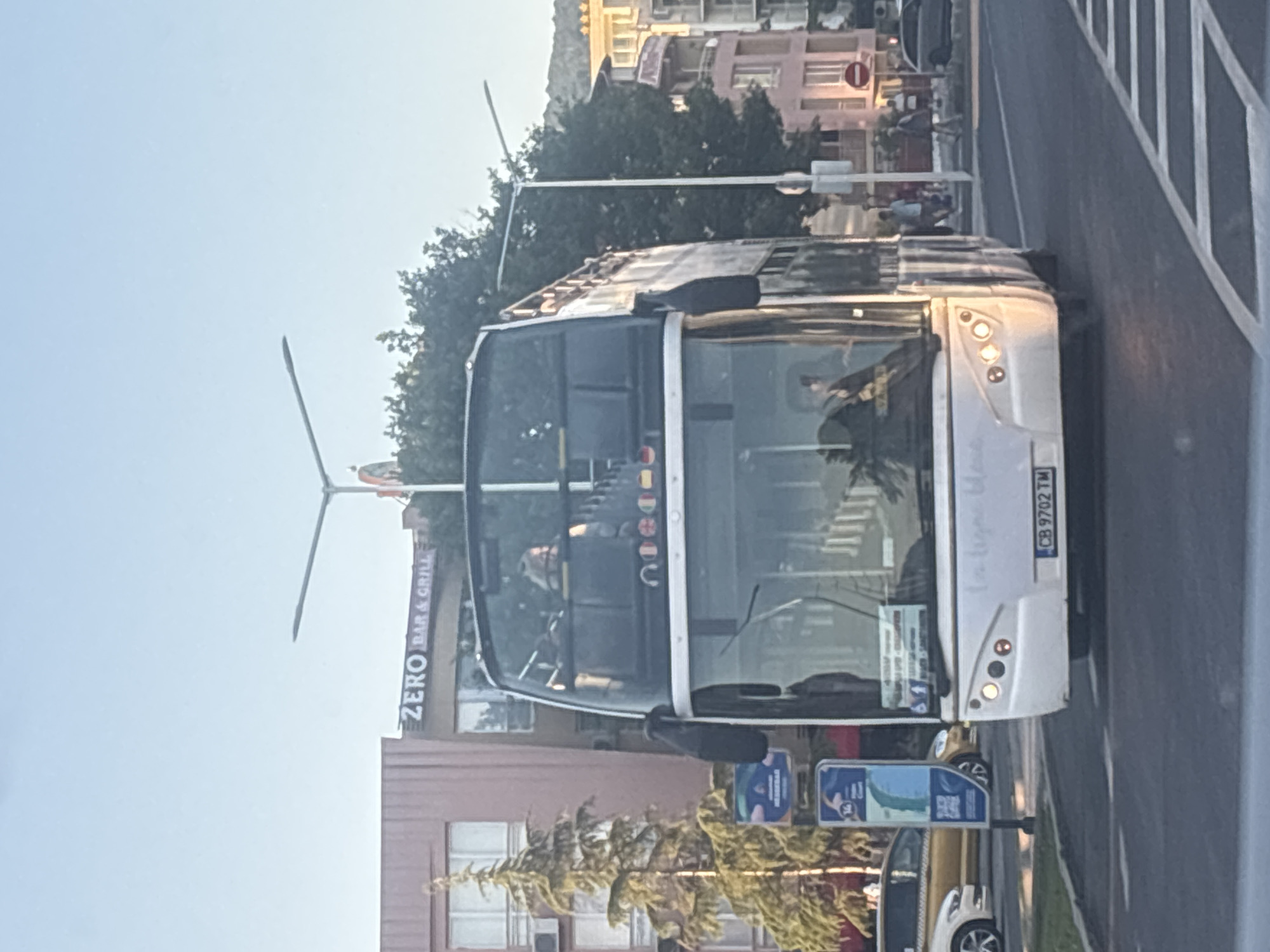 A white double-decker bus is traveling on a street in Nesebar, Bulgaria. The bus has a license plate reading "CB 9702 TM". On the upper deck, passengers can be seen through the windows. In the background, there is a building with a sign that reads "ZERO BAR & GRILL". Several trees and streetlights are also visible. Other vehicles and pedestrians are present on the street. The sky is clear and blue, indicating a sunny day.