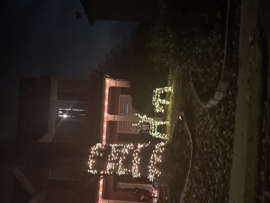 A house is decorated with Christmas lights. Two light-up reindeer stand on the front lawn. A string of lights spells out "CHEER" vertically along the front of the house. The house has columns and red brick, and a window is visible on the second floor. It appears to be nighttime. Bushes and landscaping surround the house and decorations.