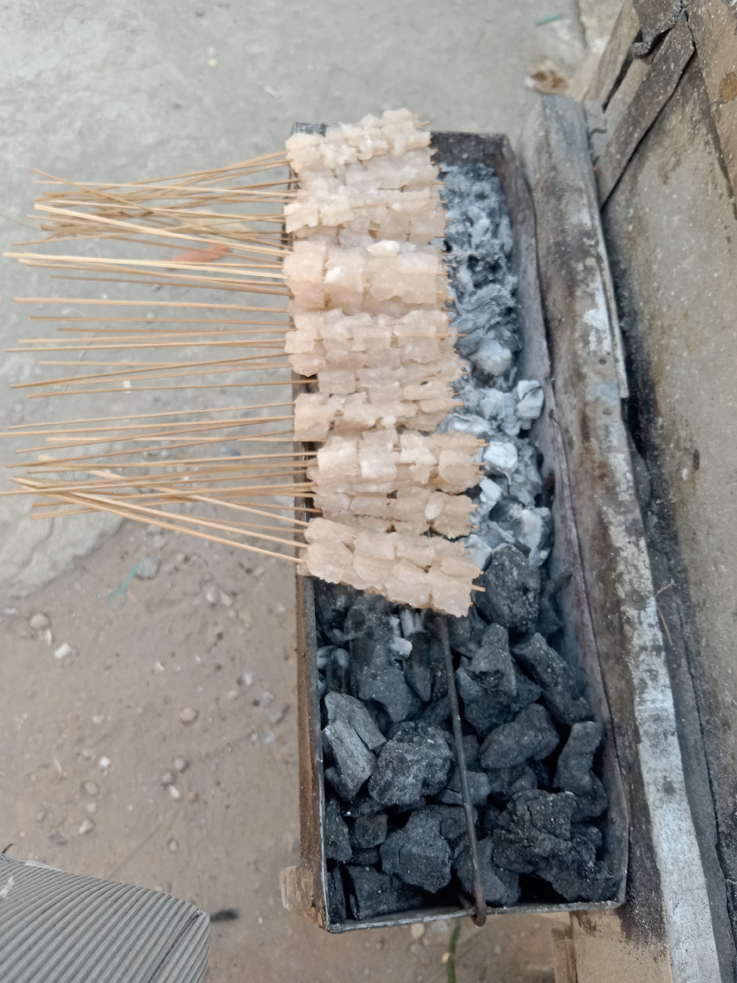 A close-up, overhead shot shows skewers of white, cubed food being grilled over hot coals. The food, possibly meat or a meat substitute, is cut into small, uniform cubes and threaded onto thin wooden skewers. The skewers are arranged in neat rows over a metal charcoal grill, the coals glowing red and emitting heat. The background is a rough, grey surface, possibly concrete or asphalt, with scattered debris. The scene appears to be outdoors, as suggested by the natural lighting and the dusty ground.