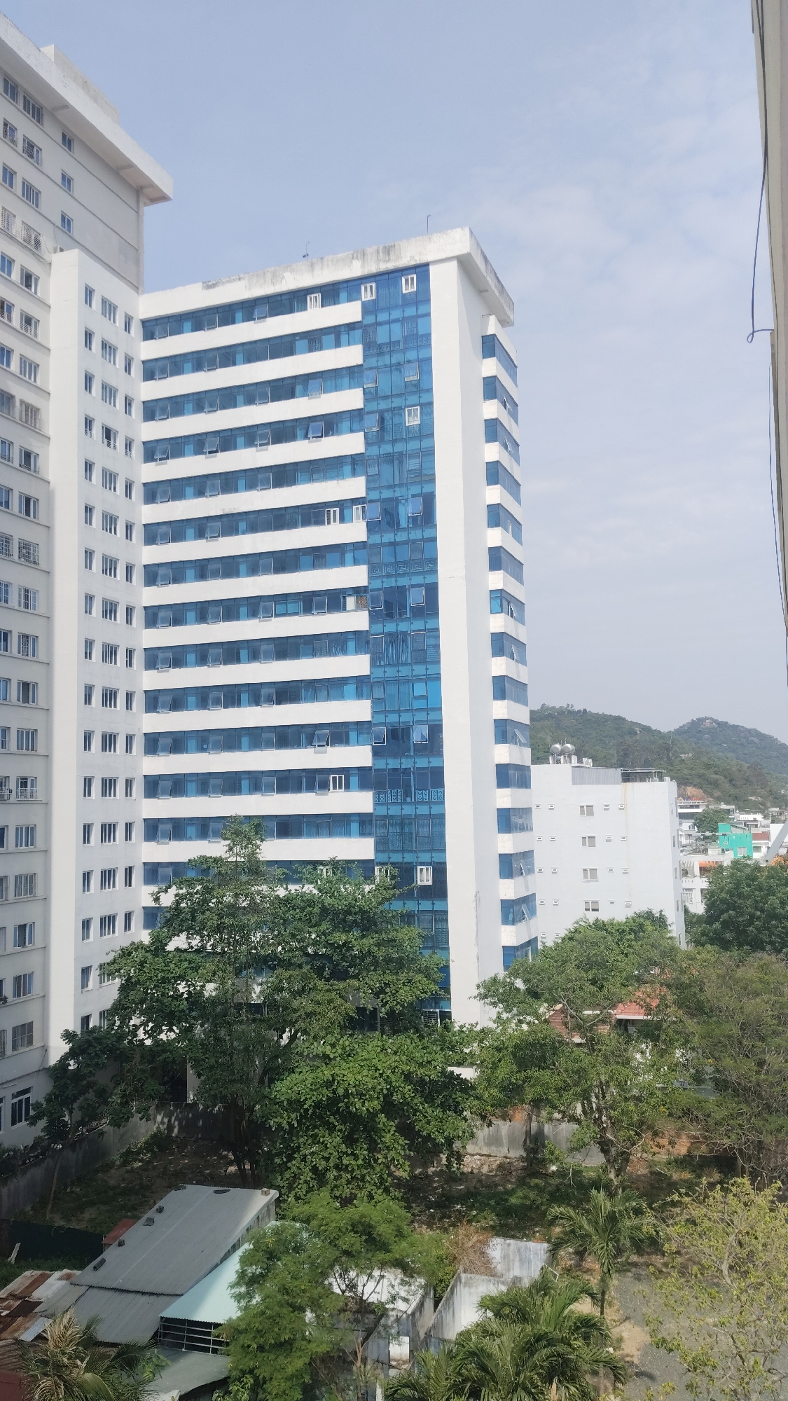 A daytime, eye-level shot shows a view from a height looking towards buildings and greenery in Bac Nha Trang Ward, Vietnam. Dominating the frame are two tall, white buildings, both rectilinear in shape. The first building on the left features rows of uniform windows running vertically along its facade. Adjacent to it, a second building has a large section with blue-tinted windows. Both appear to be mid-rise or high-rise structures.
At the base of these buildings is dense greenery, mostly leafy trees and shrubs. Several small, low-lying buildings are visible beneath the trees, with roofs of corrugated metal or tiles.
In the distance, a sloping hill can be seen, and the overcast sky has a washed-out blue color. The overall composition focuses on the contrast between the urban architecture and the surrounding nature. A vertical white line is visible in the extreme upper-right side of the image.