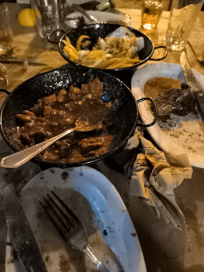 The image presents a close-up, high-angle view of a dining table laden with dishes and food remnants, suggesting a recently concluded or ongoing meal in a casual setting, likely in Nea Erithrea, Greece. The warm, dim lighting indicates it is likely evening or nighttime.
In the foreground, a black, rustic-style pan holds a dark, rich, stew-like dish with visible chunks of meat or vegetables, and a metal spoon resting within it. Behind this, another similar black pan contains strips of yellow and white food, possibly sautéed peppers and onions.
The table is cluttered with used plates and cutlery. A white oval plate to the right shows remnants of a meal, including a piece of cooked meat and sauce, accompanied by a knife. Another partially visible, dirty plate with a fork and knife lies in the lower left. Crumpled brown paper napkins are scattered on the wooden table surface.
In the background, several clear drinking glasses are visible, one containing an amber-colored liquid. A yellow object, possibly a lemon, is also present. No people or visible text are discernible. The scene conveys a relaxed, convivial atmosphere after a shared meal.