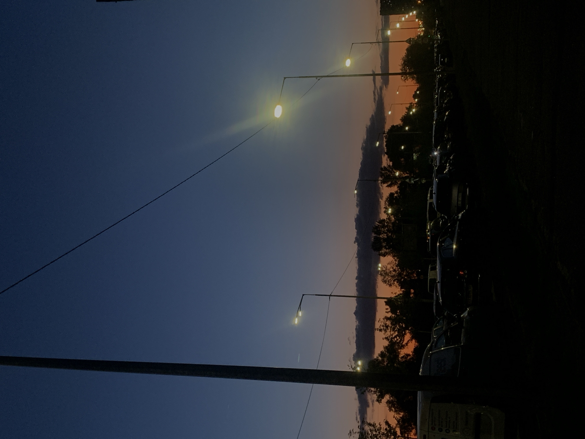 The image depicts an outdoor parking lot at dusk, likely in Kyiv, Ukraine. The sky transitions from a deep blue at the top to a warm orange and pink hue near the horizon, with dark clouds streaking across the middle ground. Several streetlights are illuminated, casting a bright glow and creating lens flares. The foreground is dominated by the dark silhouettes of parked cars and a large utility pole on the left. Trees are visible behind the parked vehicles. The scene suggests a typical urban environment at the end of the day.