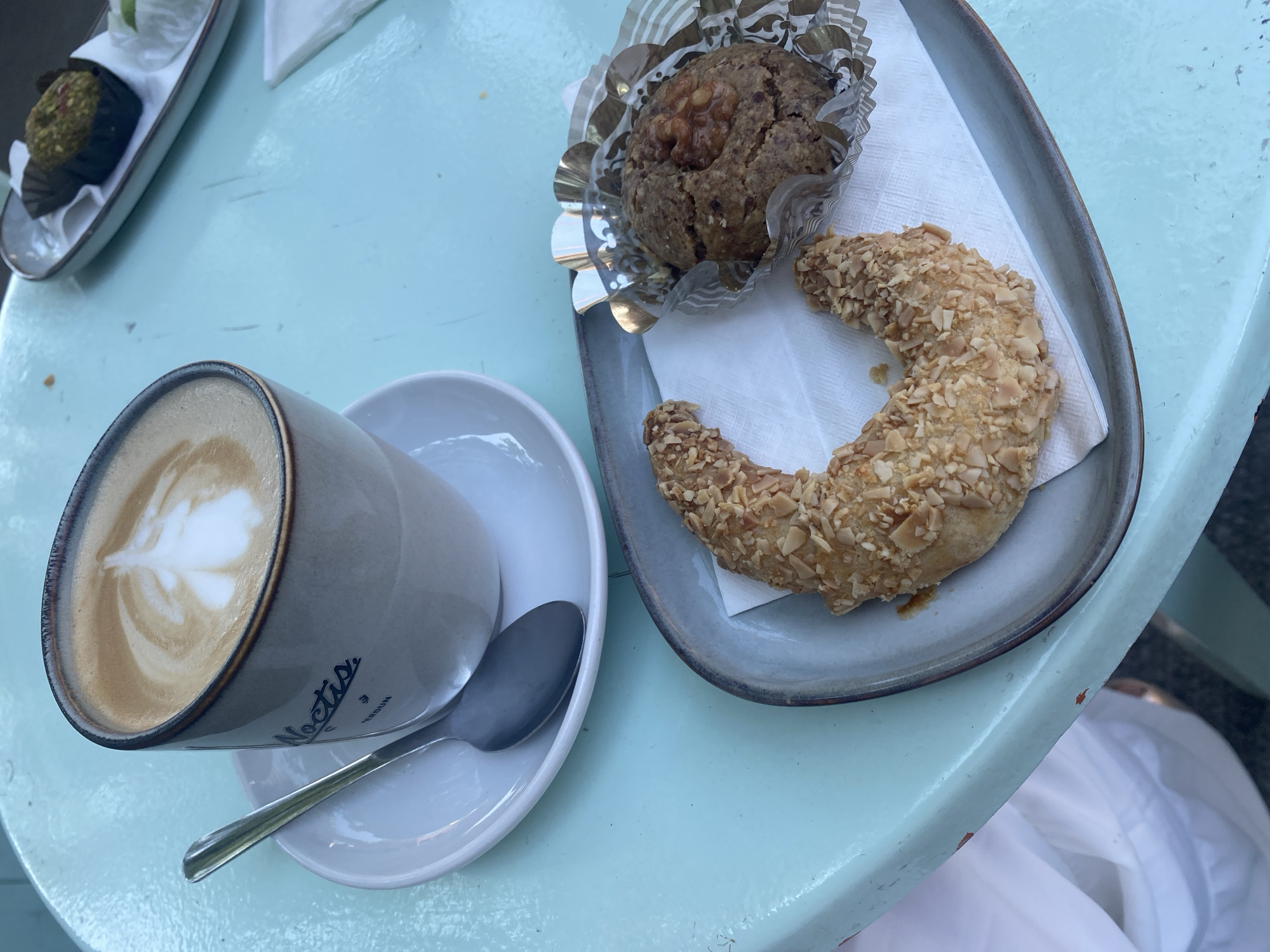 The image shows a close-up of a light blue table, suggesting an outdoor cafe setting. On the table, there is a beige ceramic mug with a latte, a white saucer, and a silver spoon. The mug has the word "Noctis" and "VERDUN" printed on it in black lettering. To the right of the mug, a grey oval plate holds a crescent-shaped pastry covered in chopped nuts and a small round baked good topped with a walnut. The pastry appears to be a cornet or croissant, while the round baked good resembles a cookie or small cake. In the background, another plate with a small green item and some crumbs is visible. A white napkin is placed under the pastries. The overall lighting suggests it is daytime. The presence of "VERDUN" on the mug indicates the cafe is likely located in the Verdun borough of Montreal, Canada.