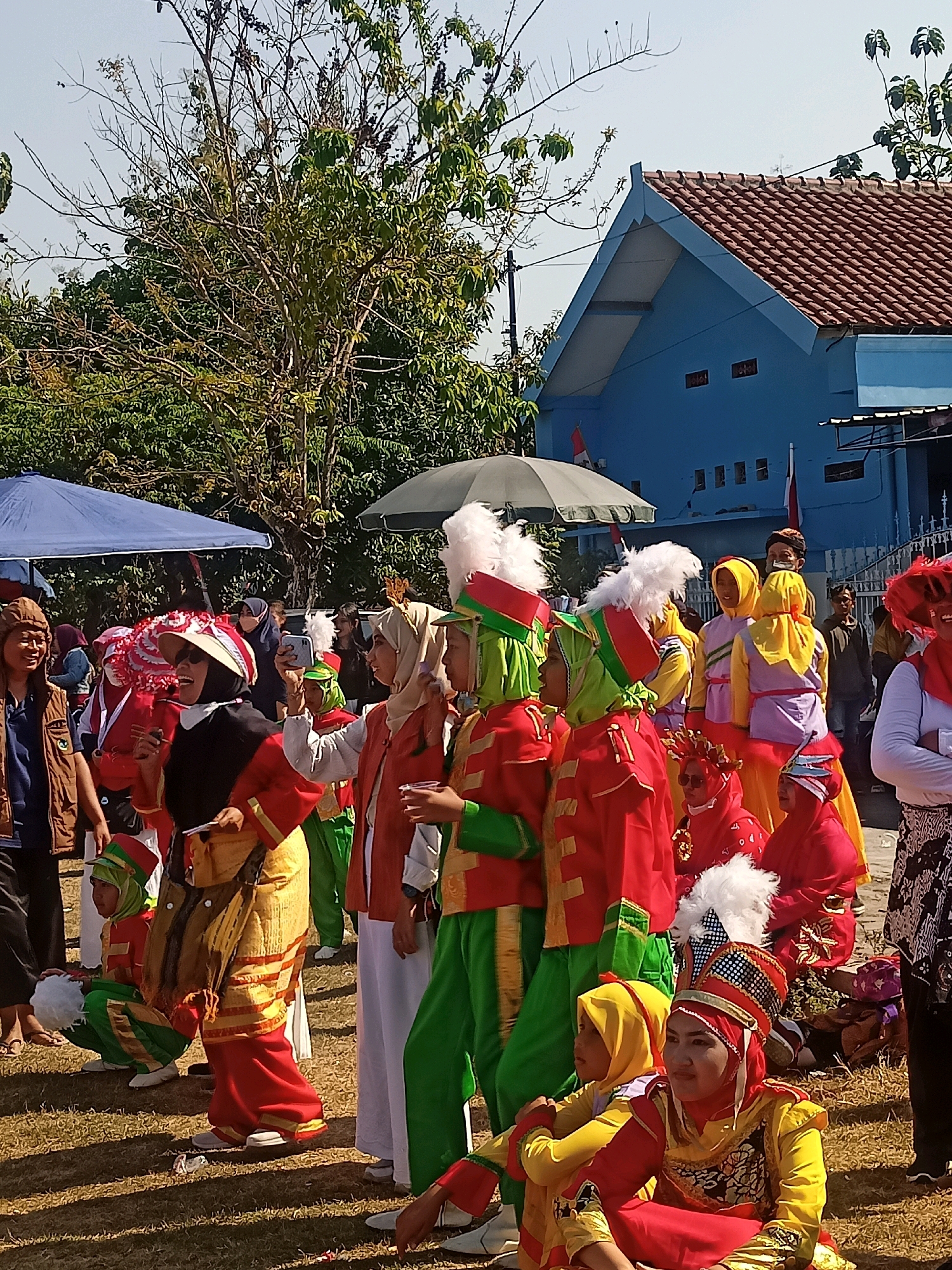 In Sumberlawang, Indonesia, a group of children and adults are gathered outdoors, participating in what appears to be a parade or cultural performance. The participants are dressed in vibrant, traditional costumes. Many of the costumes feature bright red and yellow fabrics, some adorned with gold trim and intricate patterns. Several individuals wear tall, feathered headdresses, adding to the festive atmosphere. One child in the foreground is sitting on the grass, wearing a yellow hijab and a red and gold costume, looking towards the left. Another child, also seated, is in a green and red outfit with a green hijab. Adults in the background are also dressed in traditional attire, with some holding phones, possibly recording the event. A blue building with a red tiled roof is visible in the background, suggesting a public space or community area. The ground is covered in dry grass, indicating an outdoor setting in a possibly tropical climate. Trees with sparse foliage are present, and a blue tent is set up on the left side of the image. The scene is bathed in bright sunlight, casting long shadows on the ground.