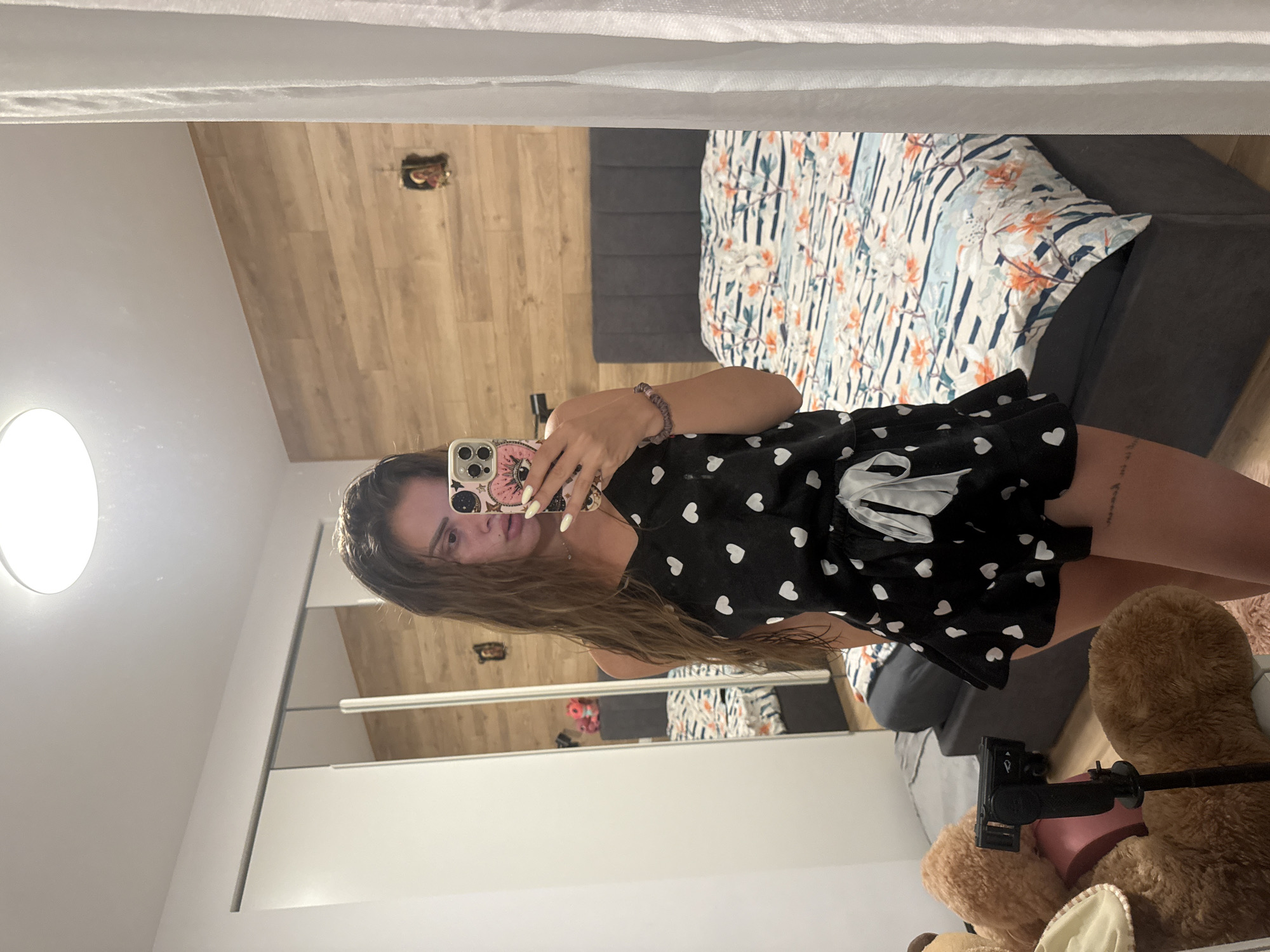 A young woman with long, wavy brown hair is taking a mirror selfie in a bedroom. She is wearing a black pajama set with white hearts printed on it. The set includes a camisole with thin straps and a pair of shorts with a ruffled hem and a white tie at the waist. She is holding a smartphone with a pink and gold patterned case. Her nails are painted yellow. On her left wrist, she wears a beaded bracelet. In the background, a bed with a patterned duvet cover is visible. To the left, there is a large teddy bear and a tripod with a camera. The room features wood-paneled walls and a white ceiling with a light fixture. The scene appears to be in a residential setting in the city of Plovdiv, Bulgaria.