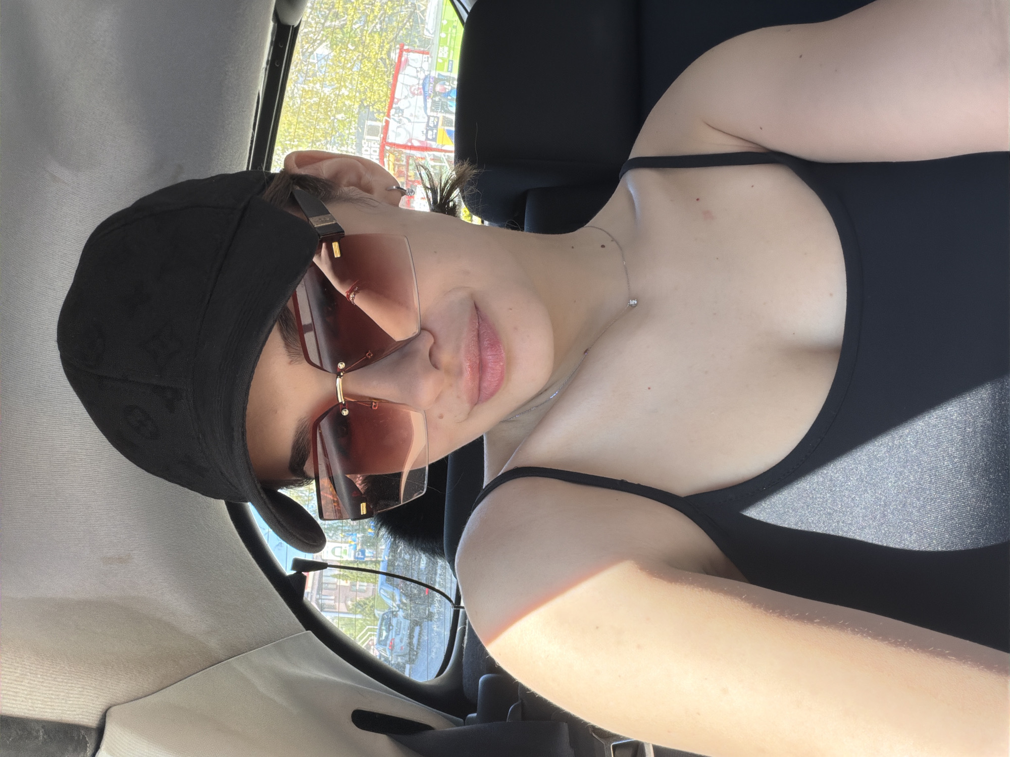 A young adult female is seated in what appears to be the front passenger seat of a car. She is wearing a black cap with a subtle patterned design and large, tinted sunglasses. Her outfit includes a black tank top with thin straps. She is wearing a delicate necklace. The background visible through the car window suggests an urban environment. Signs written in Bulgarian and other European languages are visible, leading to an inference of the location as Sofia, Bulgaria.