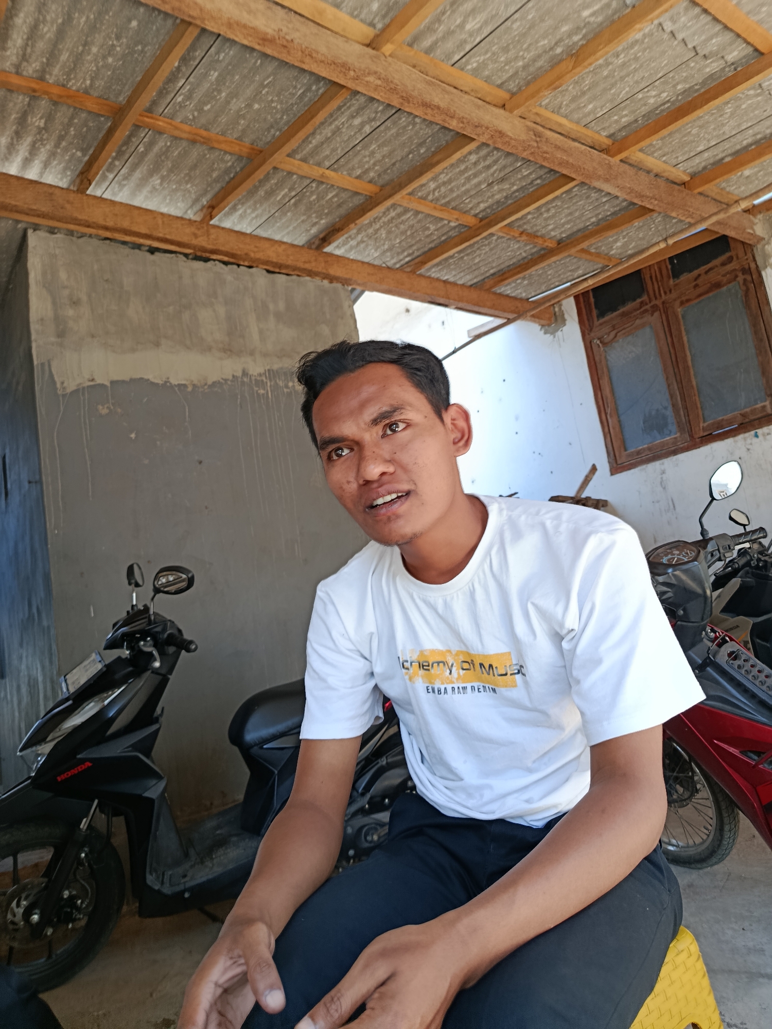 A man with dark hair and brown eyes is sitting in front of a wall in Sempol, Indonesia. He is wearing a white t-shirt with a graphic design on it and black pants. Behind him, there are two motorcycles and a wooden-framed window. The roof above is made of corrugated metal supported by wooden beams. The man is looking towards the right side of the frame, and his mouth is open as if he is speaking. The overall scene appears to be outdoors, possibly in a covered area like a carport or porch.