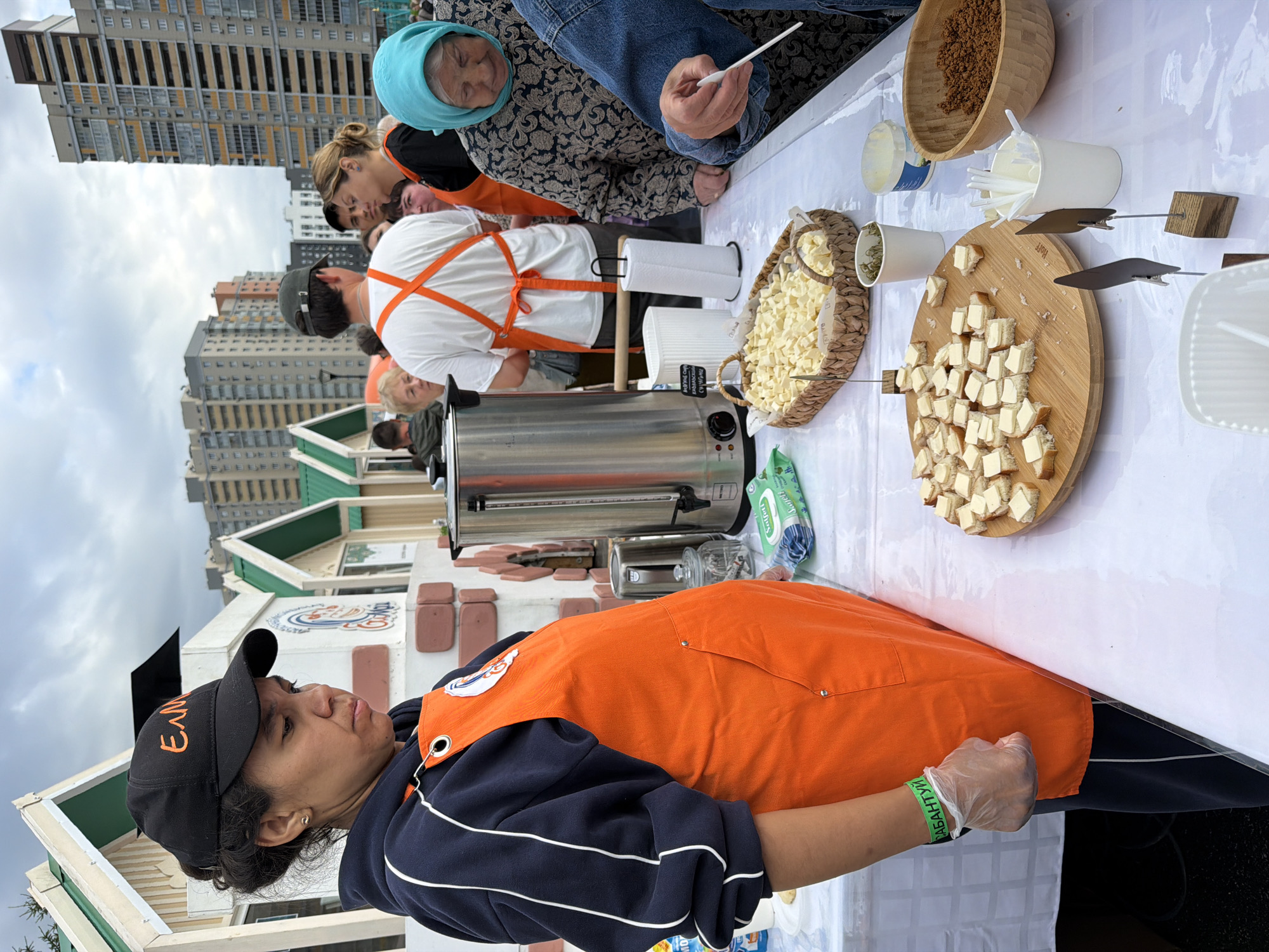In an outdoor setting with apartment buildings in the background, a woman wearing an orange apron and a black baseball cap stands behind a table laden with food. She is wearing gloves and looking down. The table displays a variety of items including a large metal thermos, a basket filled with cubed cheese, a wooden board with small sandwiches topped with cheese, a bowl of granular food, and several small cups with utensils. Other people are visible in the background, some also wearing aprons. The overall impression is of a food stall or outdoor market.