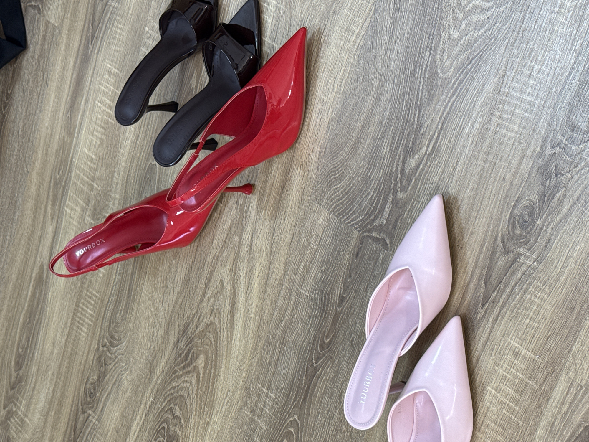 The image shows two pairs of women's high-heeled shoes on a wood-grain patterned surface. The shoes are arranged in a diagonal orientation. At the top are a pair of black patent leather mules, followed by a pair of red patent leather slingback heels. At the bottom right of the image are a pair of light pink patent leather mules. The brand name "YOURBOX" is visible on the insole of the red and pink shoes. The scene does not contain location information about Izhevsk, Russia.