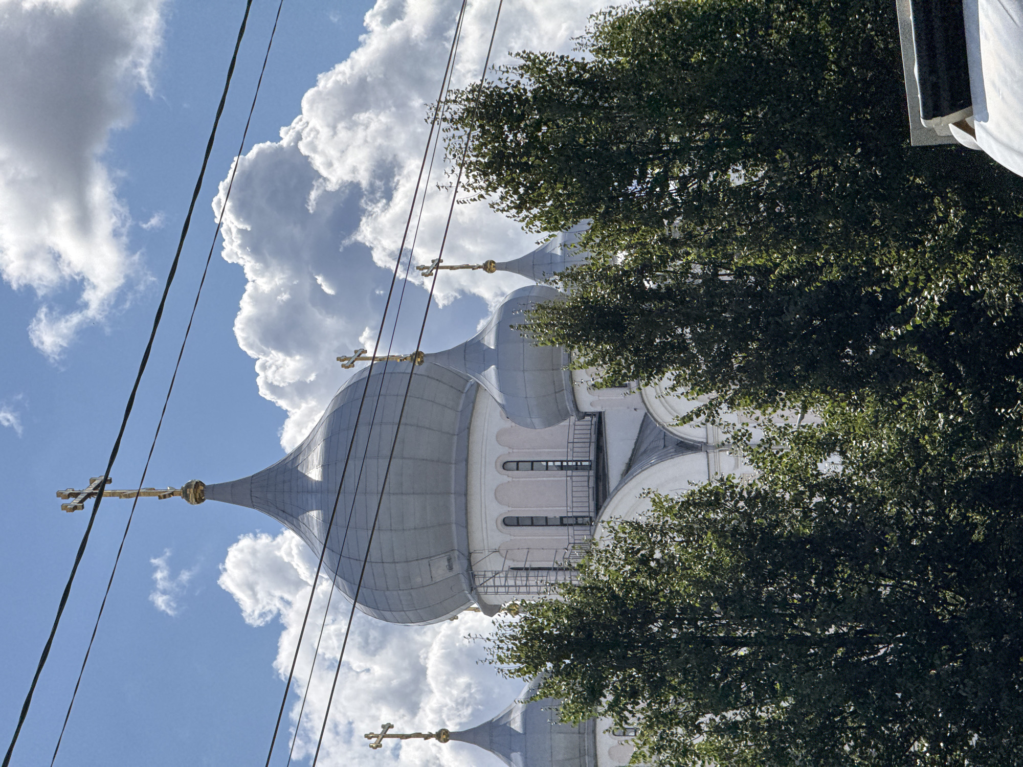 A clear, bright daytime shot of a white Orthodox church with several silver domes, two of which are topped with golden crosses, is partially obscured by the branches of green trees and thick black electrical wires strung across the frame. The church's domes are large and rounded, with one central dome being the largest. The building appears to be made of white stone or stucco, with arched windows and balconies visible. The sky is a vibrant blue, filled with fluffy white clouds that add depth to the background. The electrical wires create a grid-like pattern over the scene, with some cables running horizontally in the foreground and others diagonally. The overall impression is one of a grand, serene structure framed by nature and modern infrastructure, likely in a more urban setting within Saint Petersburg, Russia.