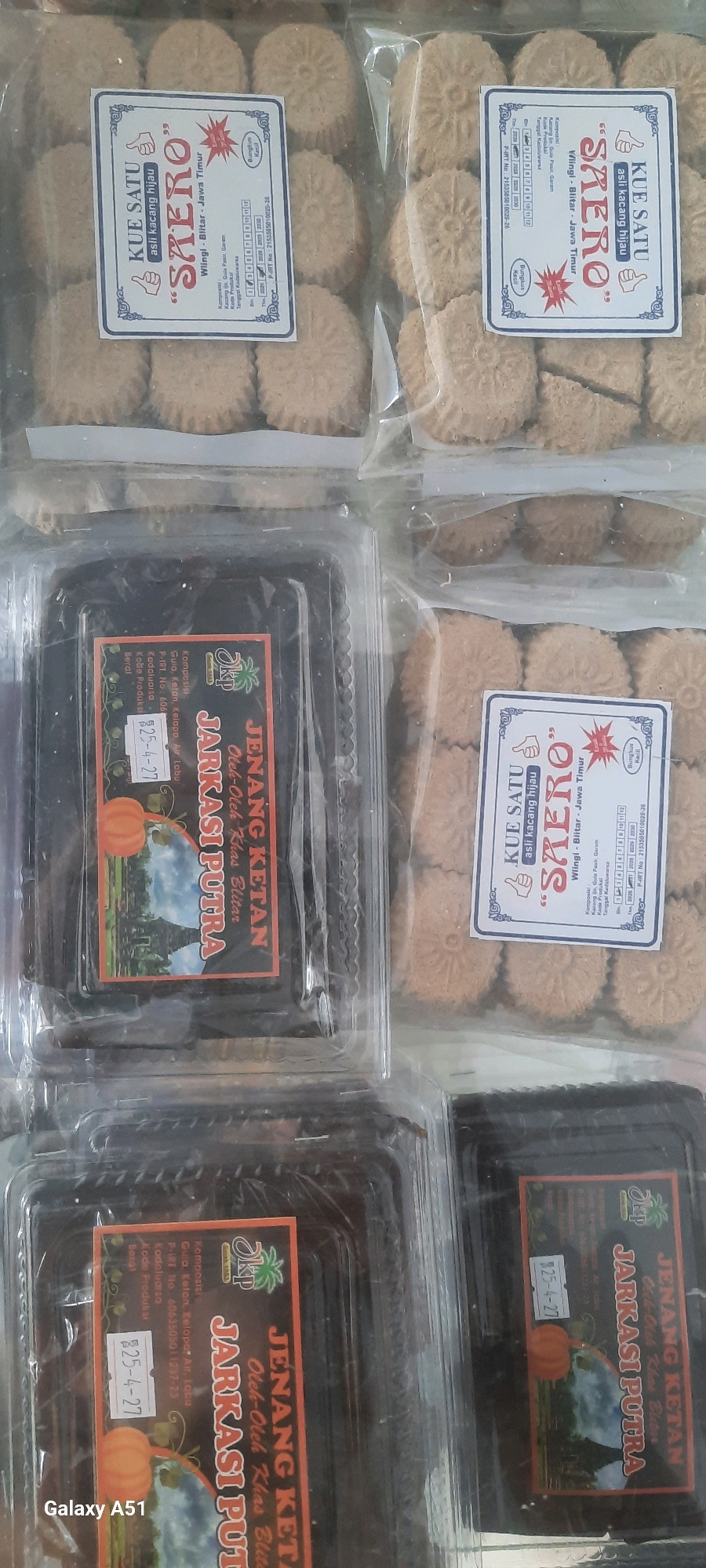 The image displays a collection of packaged snacks, likely cookies or cakes, presented in clear plastic containers. There are at least two distinct types of packaging. One type features a white label with blue and red accents, showcasing a design with a stylized floral element and text in Indonesian. The text includes "KUE SATU 'SAERO' asli kacang hijau" and indicates it is from "Wlingi - Blitar - Jawa Timur," suggesting the product originates from Wlingi, East Java, Indonesia. This packaging also includes nutritional information and a product registration number.
The second type of packaging consists of dark rectangular containers with labels that are primarily orange, black, and white. These labels also bear Indonesian text, such as "JENANG KETAN" and "Oleh-Oleh Khas Blitar," reinforcing the connection to Blitar, East Java. This packaging includes an image of a pumpkin and a temple-like structure, along with a date "25-4-27."
The snacks themselves appear to be small, round, and light brown in color, with a textured surface. They are arranged in rows within their transparent packaging, allowing for visibility of the quantity and arrangement of each product. The overall impression is of commercially produced food items, likely intended for sale. The lighting in the image is somewhat diffused, suggesting an indoor setting, possibly a retail display or storage area. The presence of a "Galaxy A51" watermark in the lower left corner indicates the image was captured using that specific mobile device.