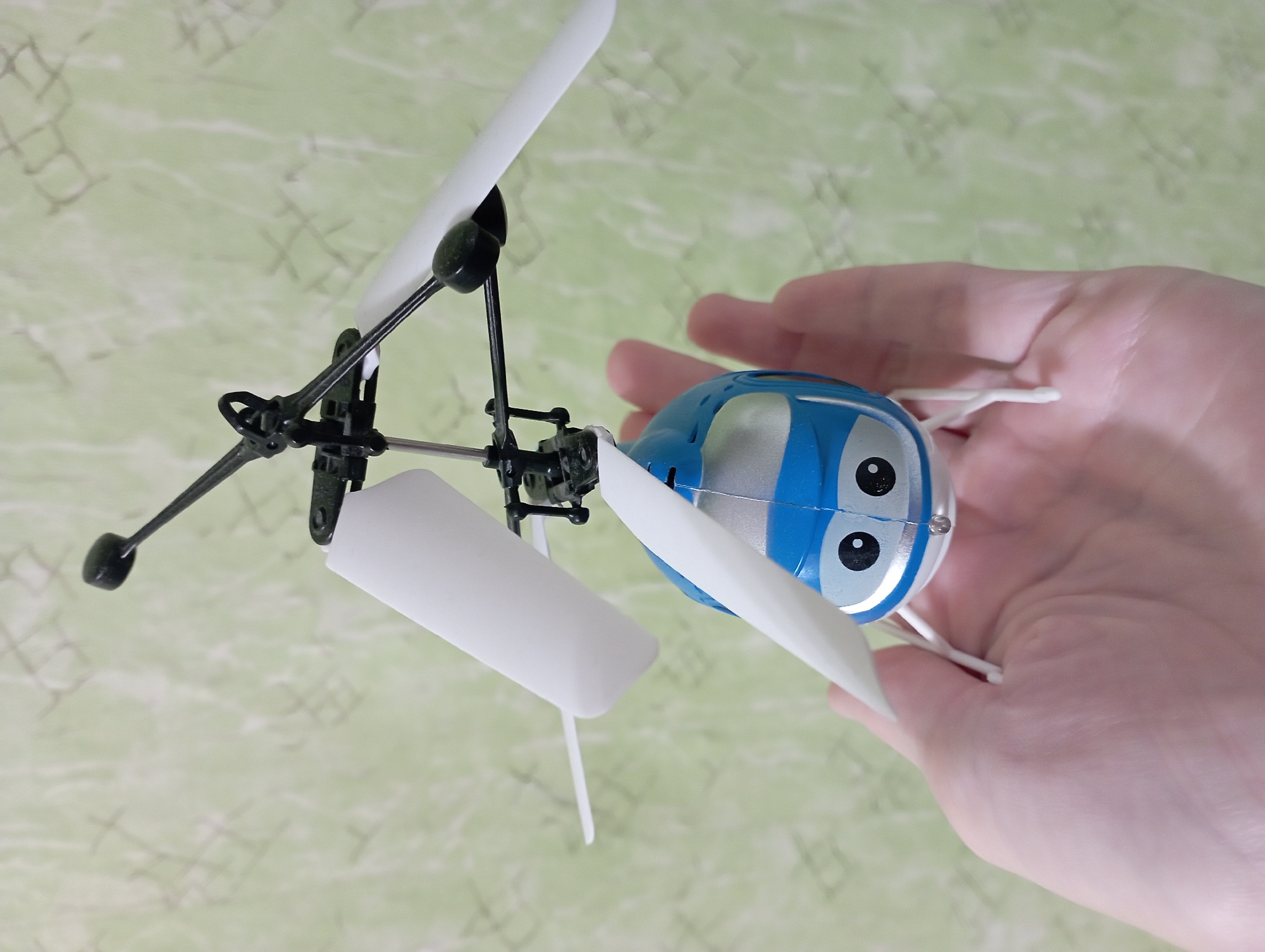 The image shows a person holding a toy helicopter. The helicopter is blue with large white eyes and silver accents, giving it a cartoonish appearance. It has two white rotor blades and a black frame. The person's hand is visible, supporting the toy. The background appears to be a light green wall with a subtle pattern. The lighting suggests it could be indoors with artificial light or outdoors on an overcast day. There is no visible text in the image. The scene is a simple close-up of a person interacting with a toy.