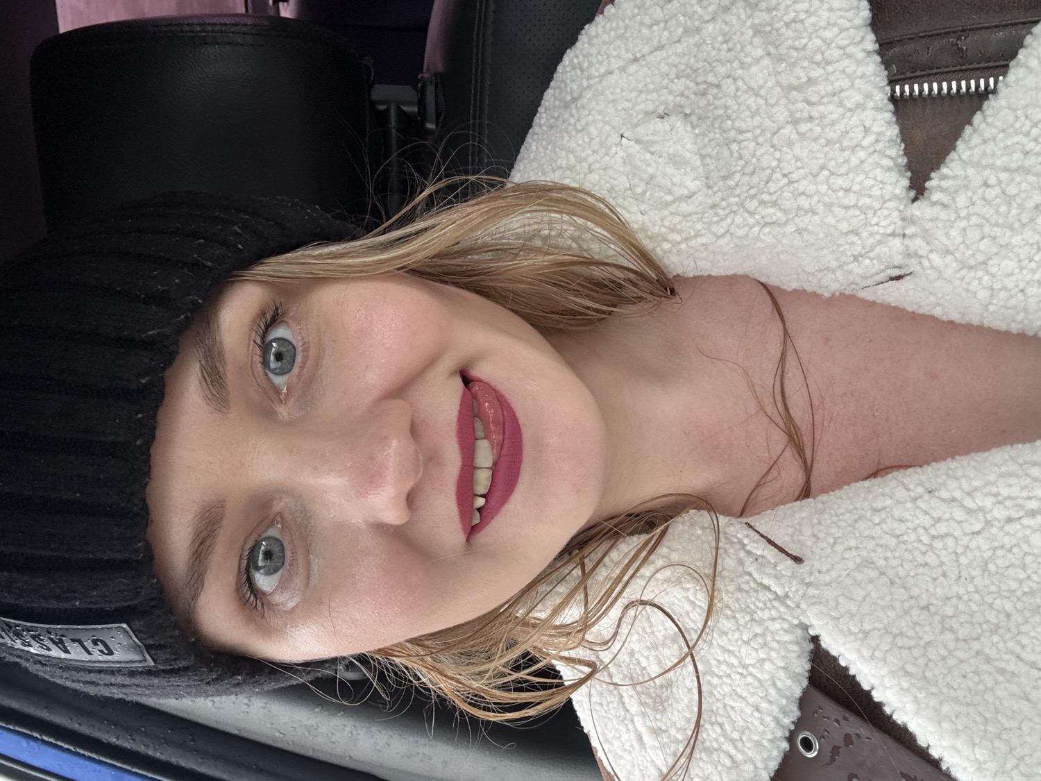A cheerful young woman is captured in a close-up selfie taken inside a vehicle in Chaikovsky, Russia. She looks directly into the camera with bright blue-grey eyes and a broad, open-mouthed smile, revealing her teeth and vibrant red-pink lipstick. Her light blonde or light brown hair appears slightly damp, with strands draped across her neck and chest, where her fair skin shows some redness.
She is wearing a cozy, white, fluffy sherpa-style jacket, which seems to be layered over a dark brown garment, possibly leather or suede, visible near her right shoulder. A black knit beanie hat covers the top of her head, featuring a rectangular patch with the text "CLASSIC" clearly visible in white. Small water droplets are scattered across her skin and the fabric of her jacket, suggesting recent exposure to rain or a humid environment. The background reveals a dimly lit car interior, primarily dark upholstery, indicating she is in a vehicle. Her overall expression is one of warmth and contentment.