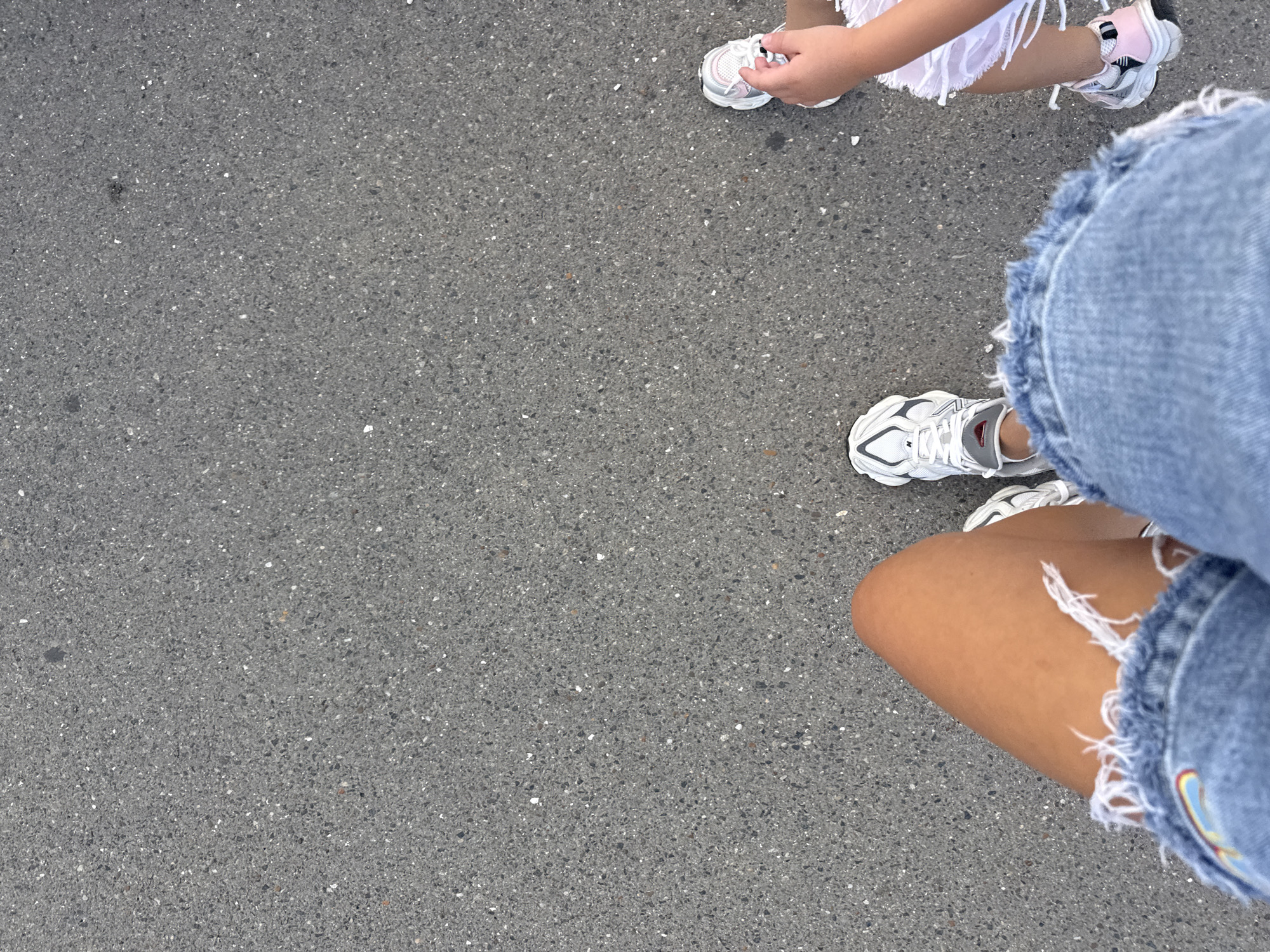 The image depicts a ground-level view of a paved surface, likely asphalt, showing two individuals' legs and feet. The person in the foreground is wearing denim shorts with frayed edges and white sneakers. Their right leg is visible from the thigh down. To the right, a smaller individual is partially visible, wearing white shorts with frayed hems and pink and white sneakers. The scene appears to be outdoors on a street or walkway, with no specific landmarks to identify it as being in Chișinău, Moldova. The lighting suggests it is daytime.
