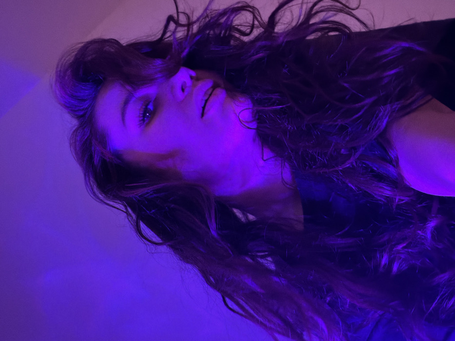 A woman with long, wavy brown hair lies on her back, facing the camera. The lighting is primarily a strong, vibrant purple, casting shadows and highlights across her face and hair. Her expression is slightly pensive, with her lips parted slightly. The camera angle is from above, giving a unique perspective of her face and hair spread out against the surface. The background is indistinct due to the strong lighting.