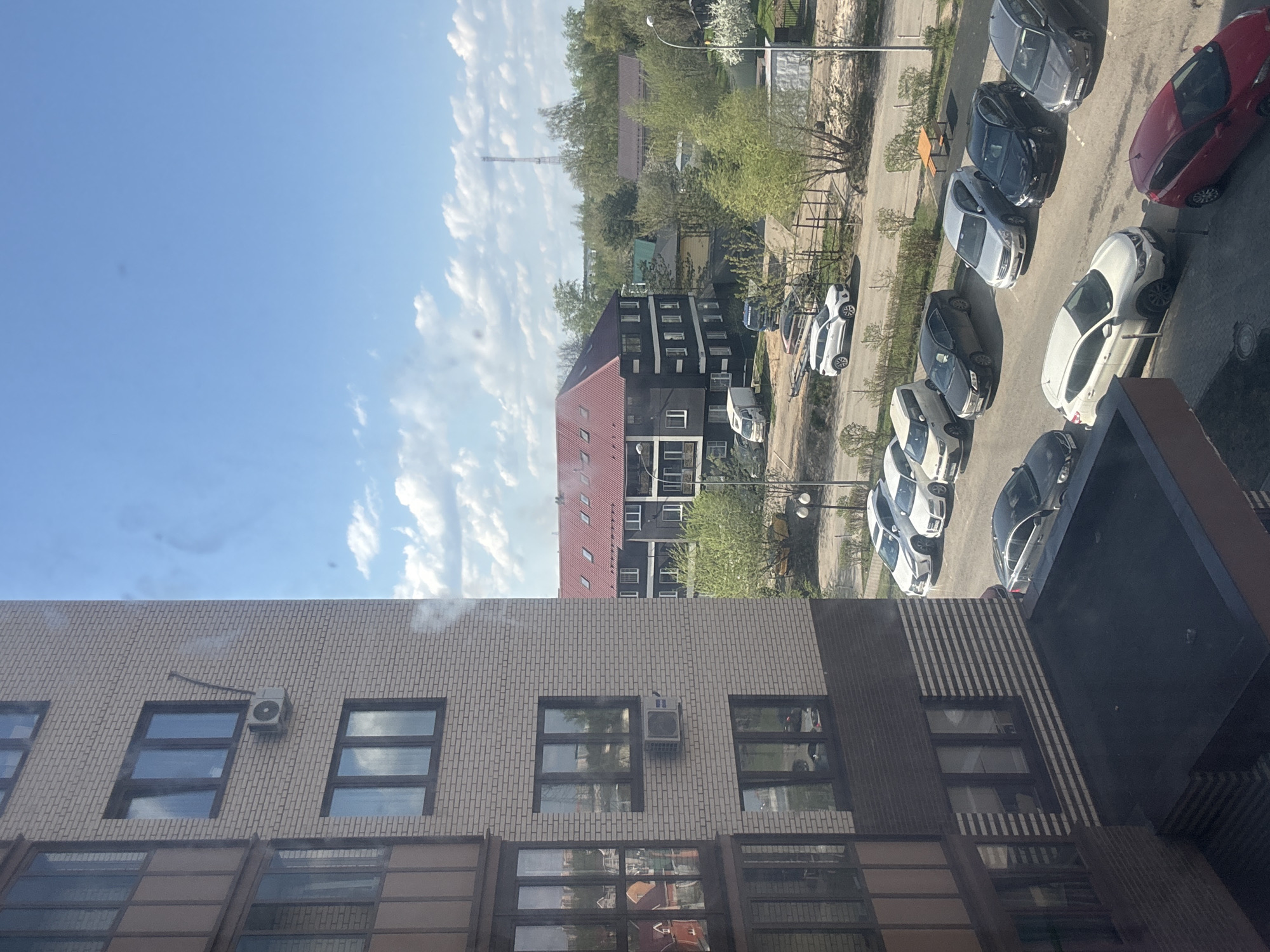 The image shows an aerial view from a building in Odintsovo, Russia. The foreground features a brick building with visible windows and air conditioning units. The background contains a mix of residential buildings, greenery, and a line of parked cars along a street. The sky is blue with scattered clouds. A smokestack-like structure is visible amongst the trees.