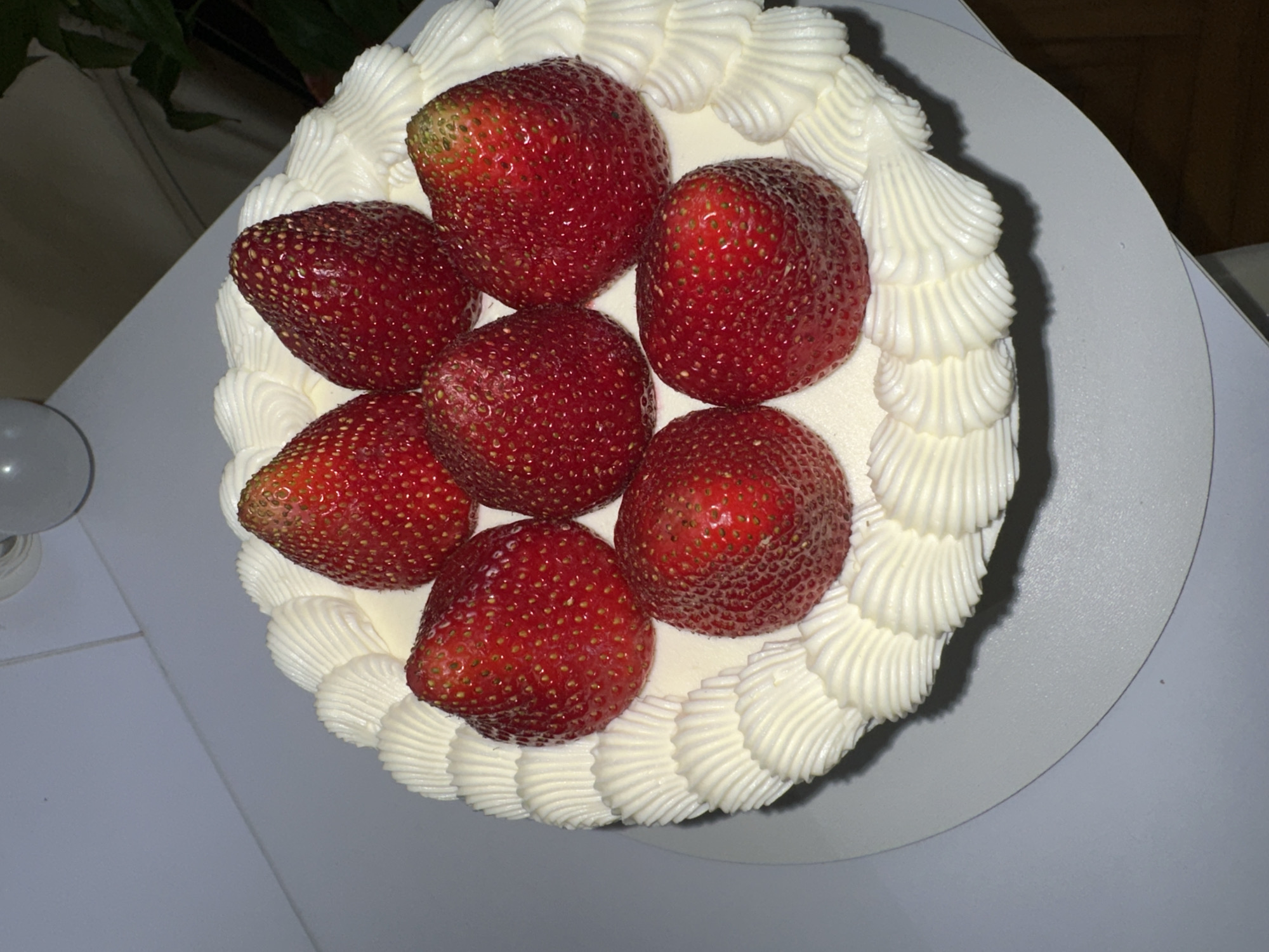 A top-down, high-angle shot shows a round cake decorated with fresh strawberries and white frosting. Six strawberries are arranged on top of the cake, appearing bright red and glossy. The frosting forms a decorative border around the strawberries. The cake sits on a white plate. The overall color scheme is dominated by red and white. Part of what appears to be a plant is visible in the upper-left corner. A lightbulb is visible at the bottom-left. The photo was taken indoors. There is no evidence to confirm the location in Ternopil, Ukraine.