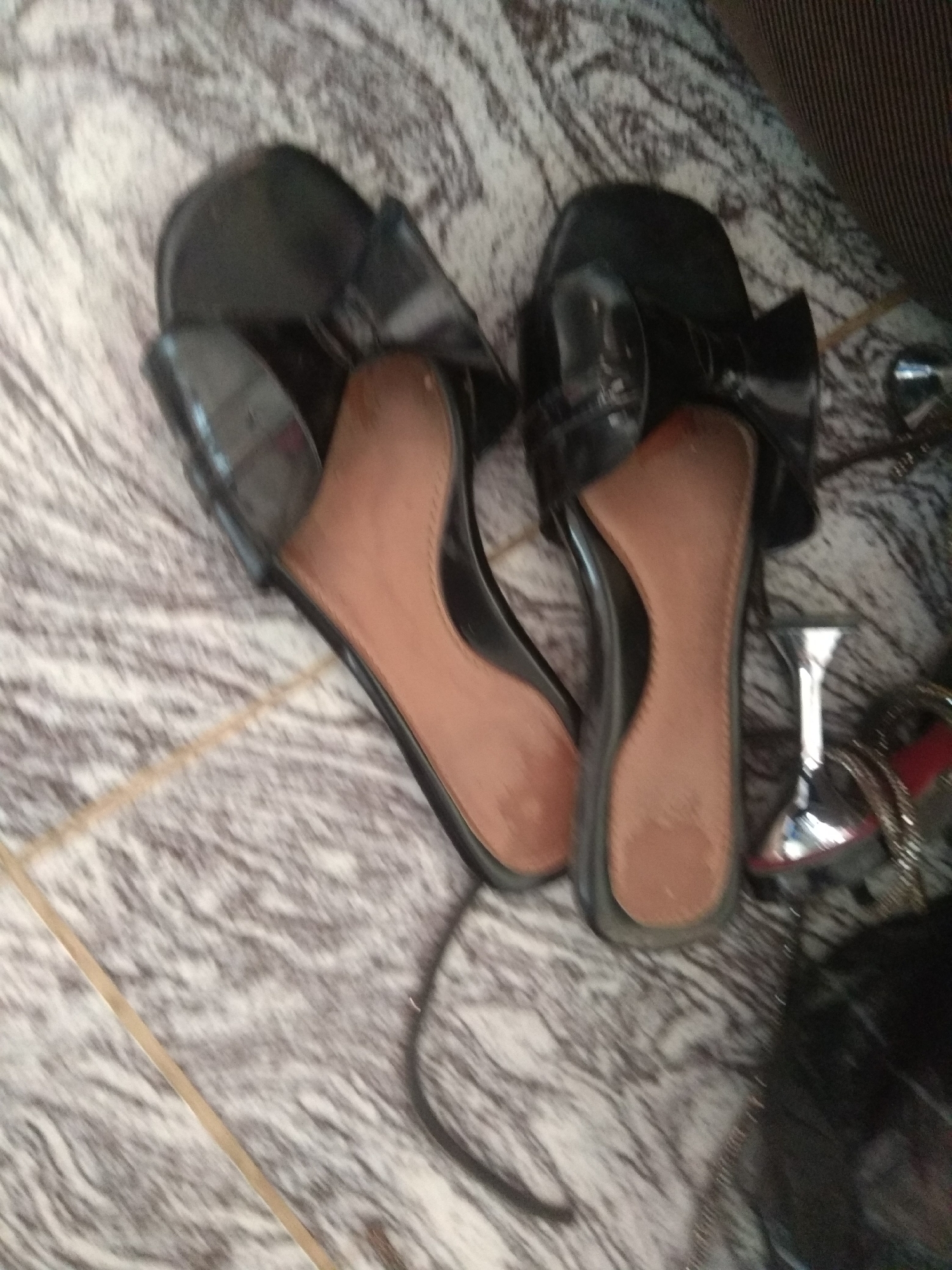 In Jalingo, Nigeria, on a marbled floor, lies a pair of black high-heeled sandals. The sandals are open-toed, with a strap across the toes and a strap around the ankle. The insoles are a light brown color. To the right of the sandals, a silver stiletto heel and a black handbag are visible. The image is taken from a high angle, looking down at the sandals and surrounding objects.
