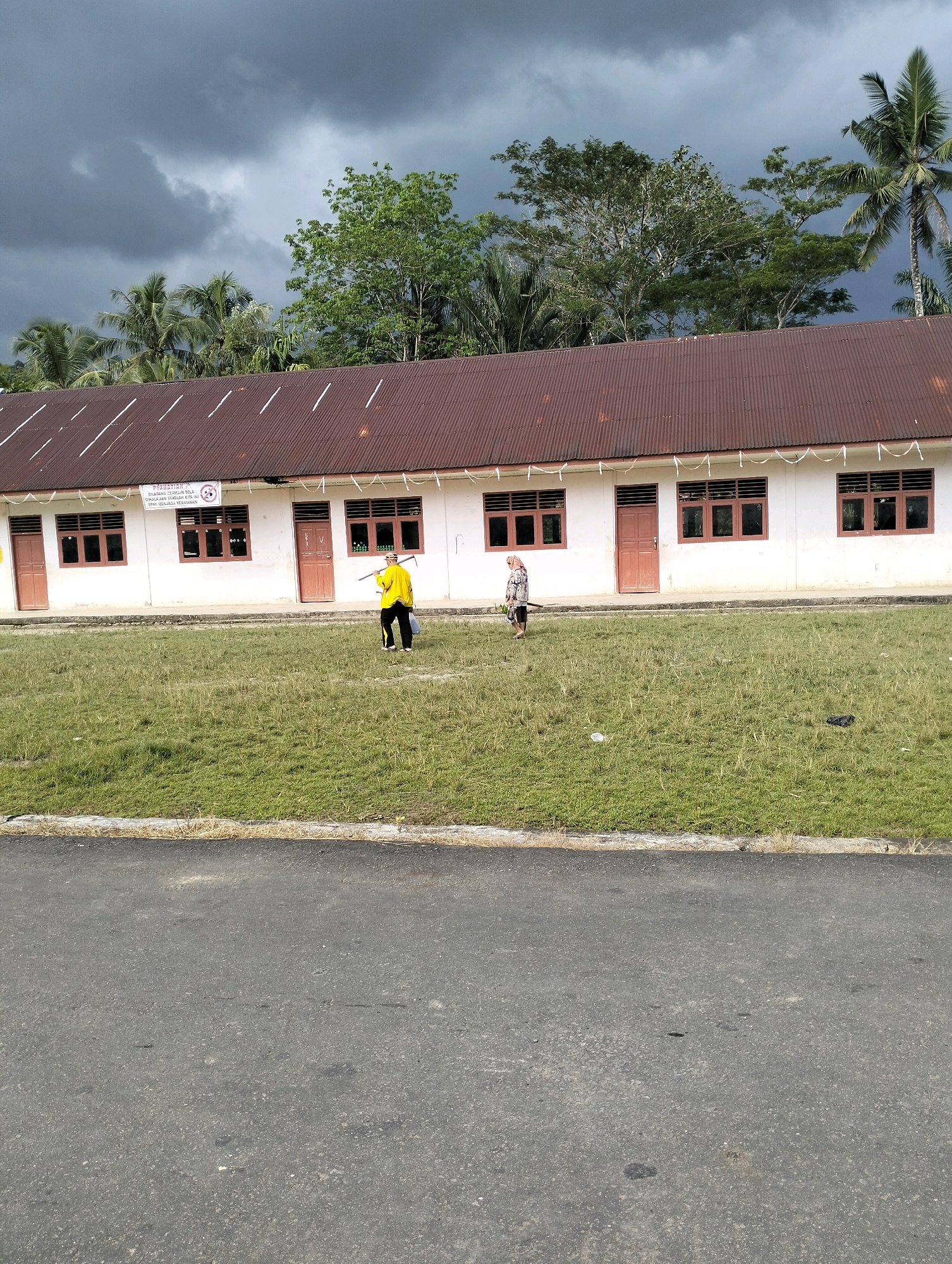 In Pekan Hilisimaetano, Indonesia, a one-story building with a corrugated metal roof and white walls stands under a cloudy sky. The building has several windows with dark frames and brown doors. In front of the building, a grassy field stretches towards the foreground where an asphalt road is visible. Two individuals are present in the grassy area. One person, wearing a yellow shirt and black pants, stands near the building, holding a long, thin object. Another person, wearing a hijab and a patterned outfit, walks away from the building towards the right side of the frame. Trees and palm trees are visible behind the building.