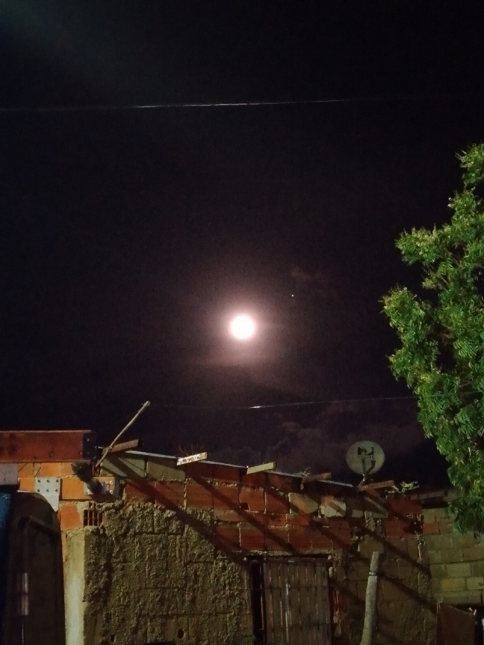 A full moon dominates a dark sky, partially obscured by wispy clouds. Below, a rustic, unfinished brick building with a satellite dish sits silhouetted against the night. The building's structure appears haphazard, with exposed brick and rough materials. A tree with dense, green foliage fills the right side of the frame. A thin, dark power line runs across the upper portion of the image.