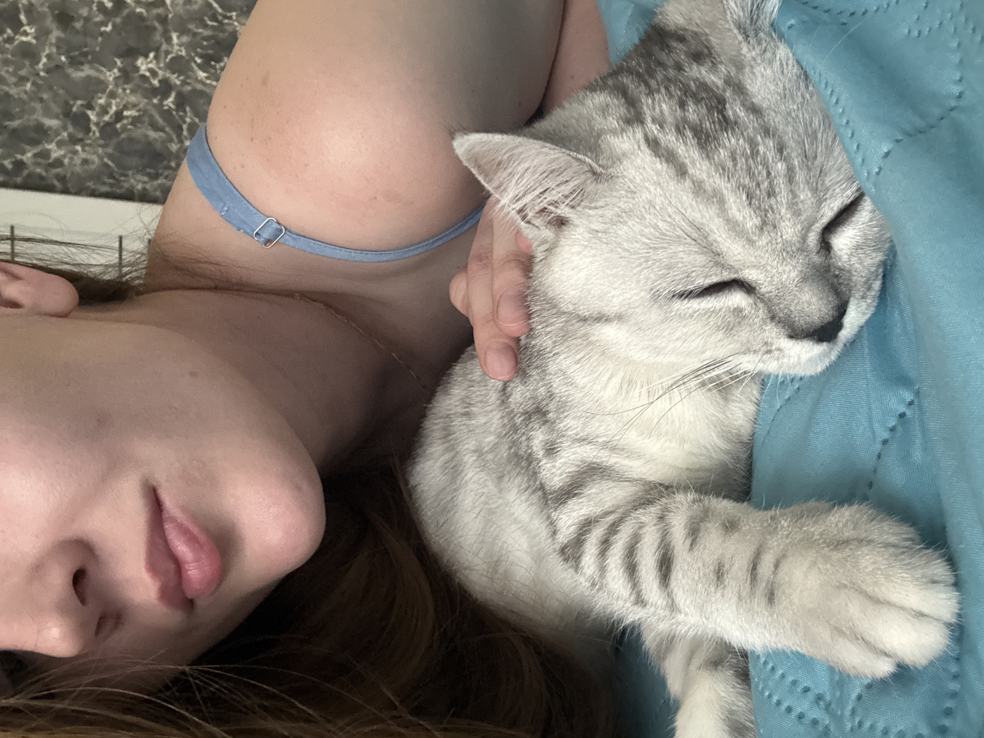 An indoor, medium-shot photograph taken in Izhevsk, Russia, depicts a woman and a sleeping domestic cat on a textured, teal-colored blanket. The woman is positioned partially on the left side of the frame, with the lower half of her face, her right shoulder, and a portion of her chest visible. Her lips are slightly parted, exhibiting a natural pink hue. Her skin is fair, and long, auburn-toned hair is visible to the left. She wears a light blue strap garment with a silver metal adjuster visible on her right shoulder. Her right hand is gently placed on the back of the cat's head. The cat is a silver tabby, displaying distinct dark grey stripes on a lighter grey base, and is observed sleeping with its eyes closed, resting on its left side. Its front left paw is visible in the lower left foreground. The background behind the woman reveals a dark grey wall with a marbled or textured pattern.