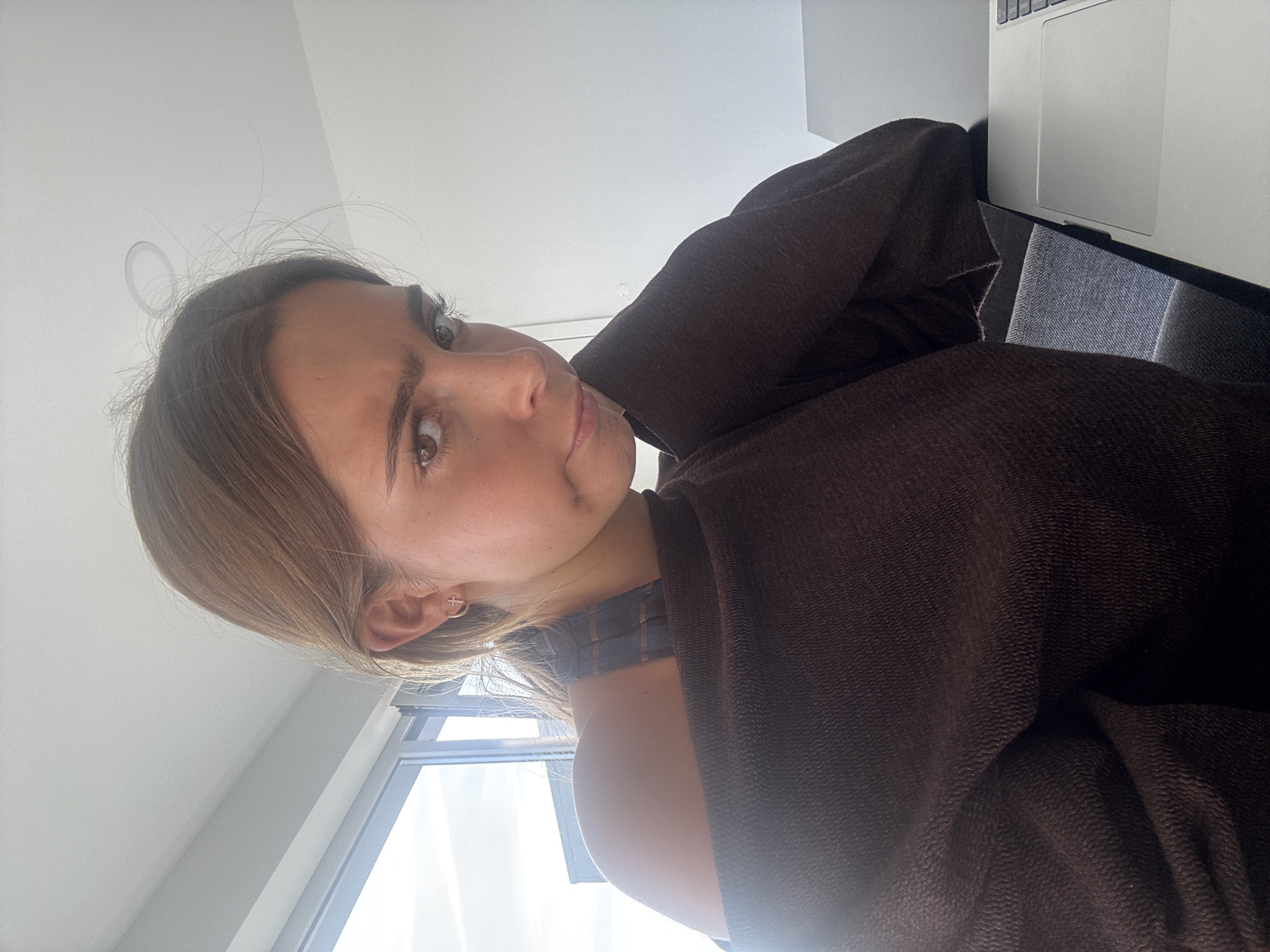 A close-up image captures a young woman looking directly towards the viewer with a calm, pensive expression, her head slightly tilted. She has light brown hair, some wisps around her forehead, and brown eyes. A small, dark mark is visible on her right cheek. She is wearing a dark brown, textured garment, possibly a robe or blanket, draped over her shoulders and right arm. A subtle plaid strap is visible underneath this garment. A delicate, cross-shaped earring is worn in her right ear.
The scene is indoors, brightly lit by natural daylight streaming through a window visible in the background to her left. The window frame is present, and the wall or ceiling above her is light-colored. To her right, a silver laptop, likely a MacBook, with its trackpad visible, rests on a dark surface next to a dark grey upholstered chair or sofa. The presence of the laptop suggests a relaxed, everyday setting, possibly a home in Torres Vedras, Portugal. The time of day is clearly daytime due to the bright natural light. No visible text is present in the image.