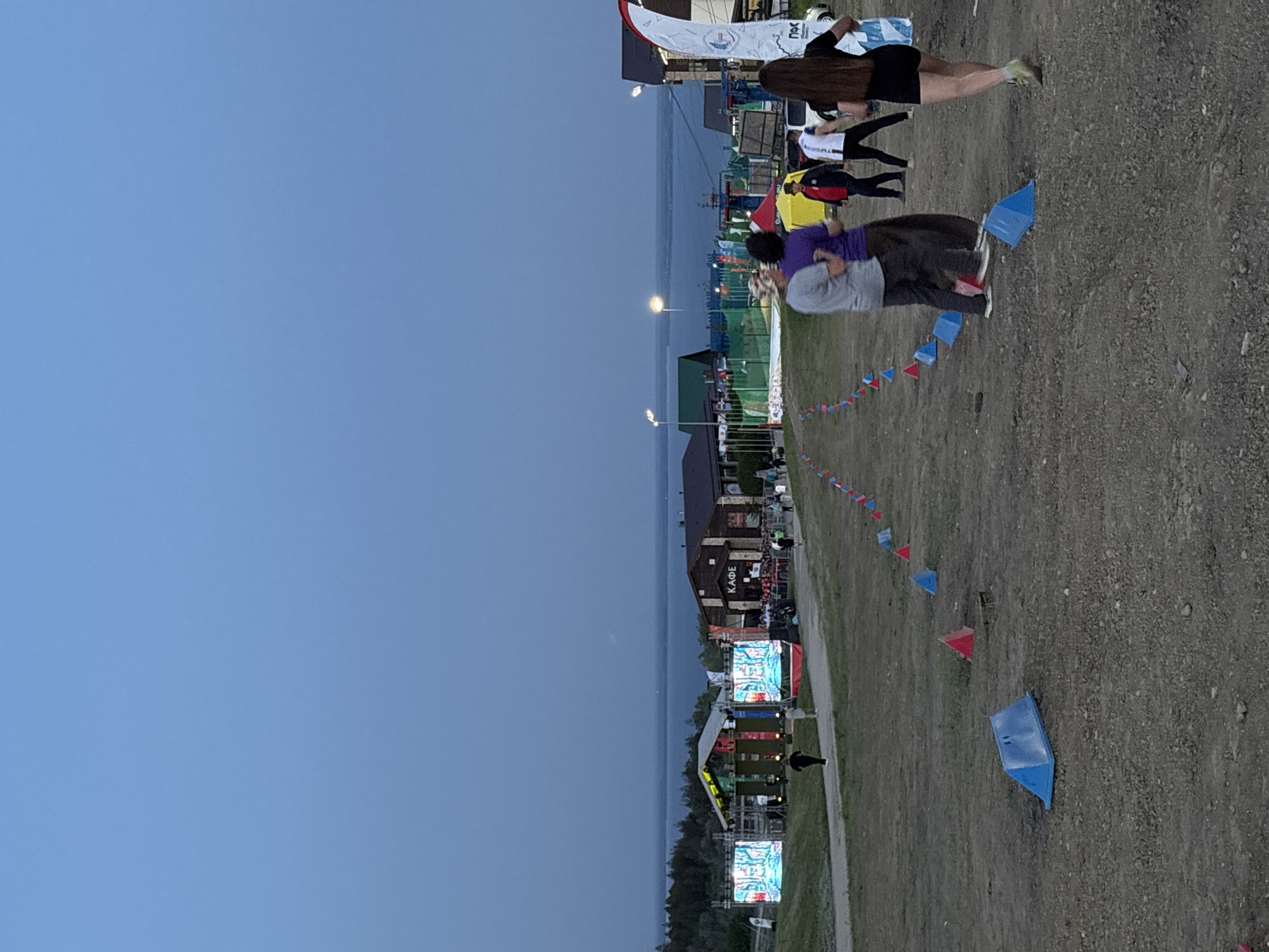 The image depicts an outdoor scene at dusk in Khvalynsk, Russia. In the foreground, a gravel path is marked with red and blue triangular flags. Several individuals are walking along the path, including a person in a purple shirt and another in a grey shirt. In the midground, there is a building labeled "KAOE" and a stage with large screens displaying images. The background consists of a body of water under a darkening sky. Streetlights are visible along the shoreline.