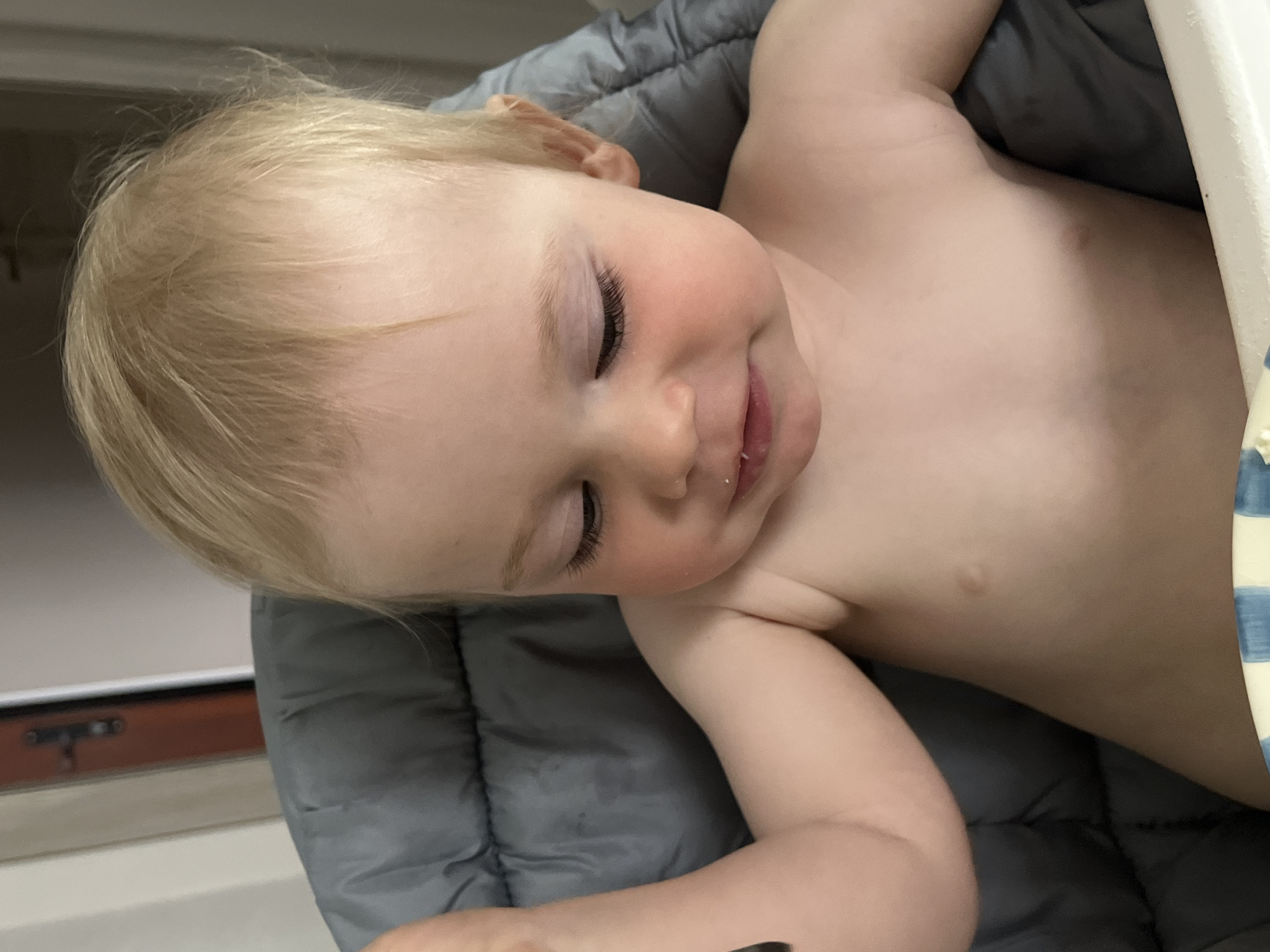 An infant with light blonde hair is seated in a grey, padded high chair. The child's bare torso is visible, revealing fair skin with subtle rosy cheeks and a small, raised blemish on the right side of the chest. The child's gaze is directed downwards and slightly to the left, with eyes partially closed, exhibiting long, dark eyelashes. A small white residue is visible on the lower lip, suggesting recent consumption of food. The left arm is slightly raised and bent at the elbow. In the background, a light-colored wall with a brown door and dark hardware is partially visible on the left side. The specific location in the city of Chaikovsky, Russia, cannot be determined from the visual information presented in the image.