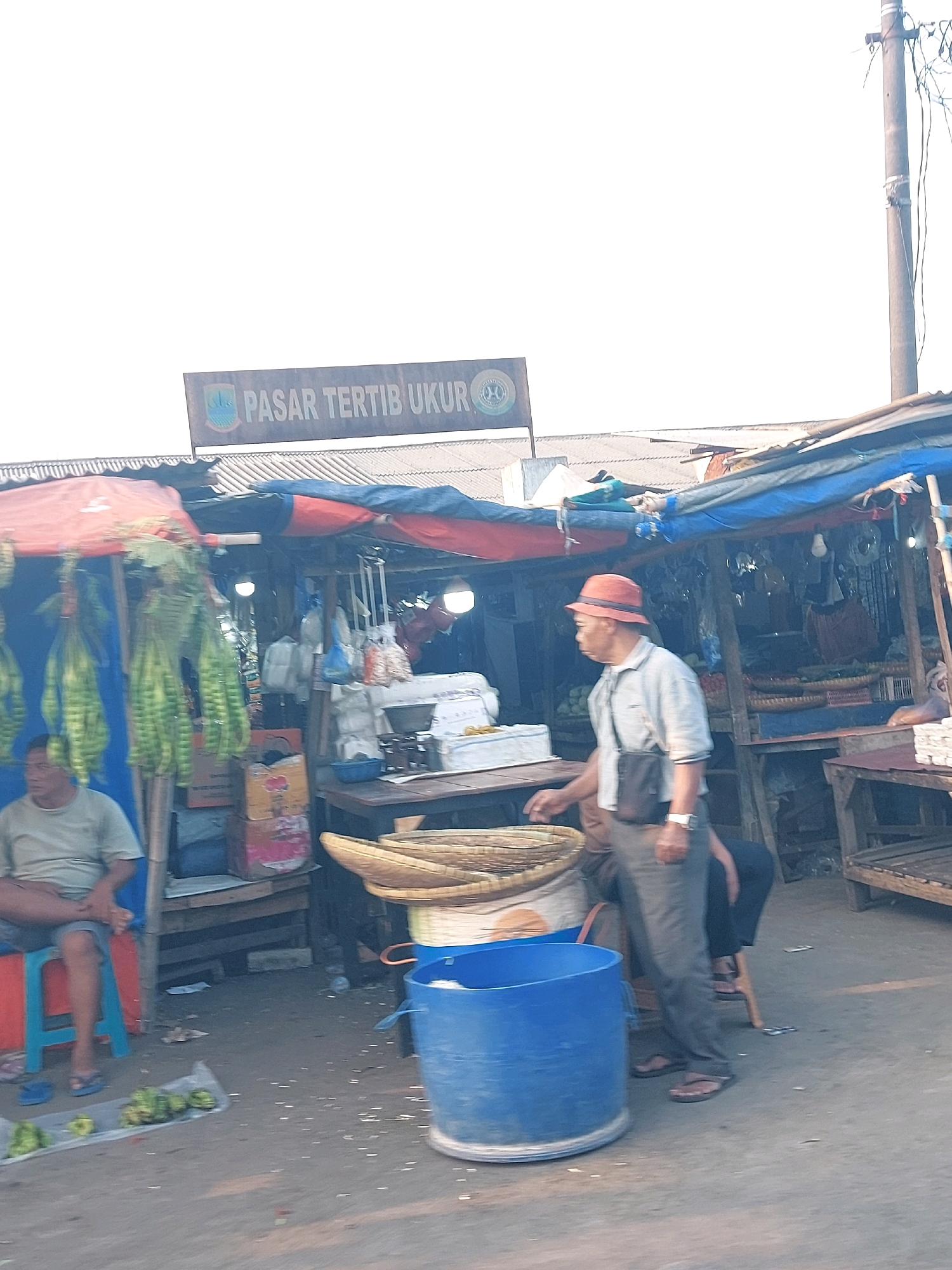 This is a photograph taken at the Pasar Tertib Ukur market in Karawang, Indonesia. The market appears to be an open-air setup with numerous stalls covered by tarpaulins, displaying a variety of goods. In the foreground, a man wearing a red hat and a light-colored shirt is standing next to a stall. He is carrying a dark bag across his body and appears to be engaged in activity related to the market. To the left of the frame, another person is seated on a low stool in front of a stall that is adorned with hanging green produce. In the background, stalls are visible with more goods, and a prominent sign that reads "PASAR TERTIB UKUR" is displayed above the market. The ground is paved, and the overall atmosphere suggests a typical bustling local market.