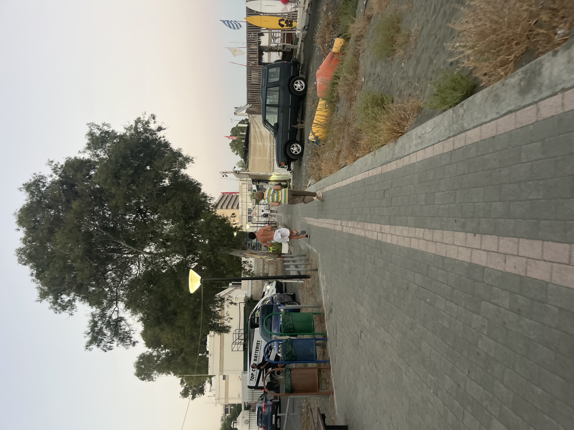 The image depicts a paved walkway in Agios Tychona Community, Cyprus, with a sandy area to the right. Two individuals are walking away from the camera on the walkway. The person on the left is a male wearing white shorts and no shirt. The person on the right is wearing a striped shirt and brown pants. In the background, several vehicles are parked, including a black SUV and vans. Trees, buildings, and streetlights are also visible. The sky is overcast, suggesting it is either early morning or late evening. There are also several buoys and surfboards on the sand.