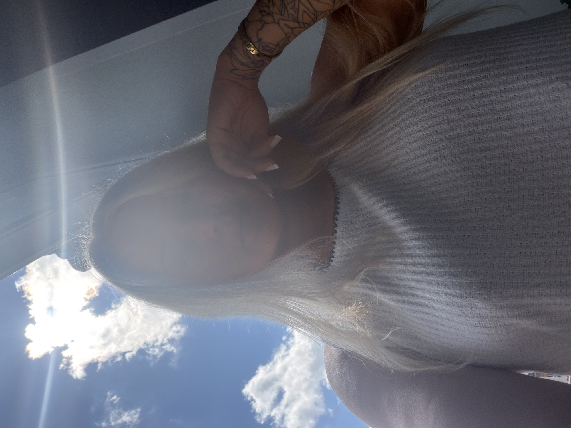 A young woman with blonde hair and tattoos on her arm poses for a selfie outdoors. She is wearing a light-colored, sleeveless top with a textured knit pattern. The sun is bright, creating a lens flare that partially obscures her face. The background shows a blue sky with white clouds and a dark structure to the right. The image does not provide any information that would allow for its location in the city of Sarapul, Russia to be determined.