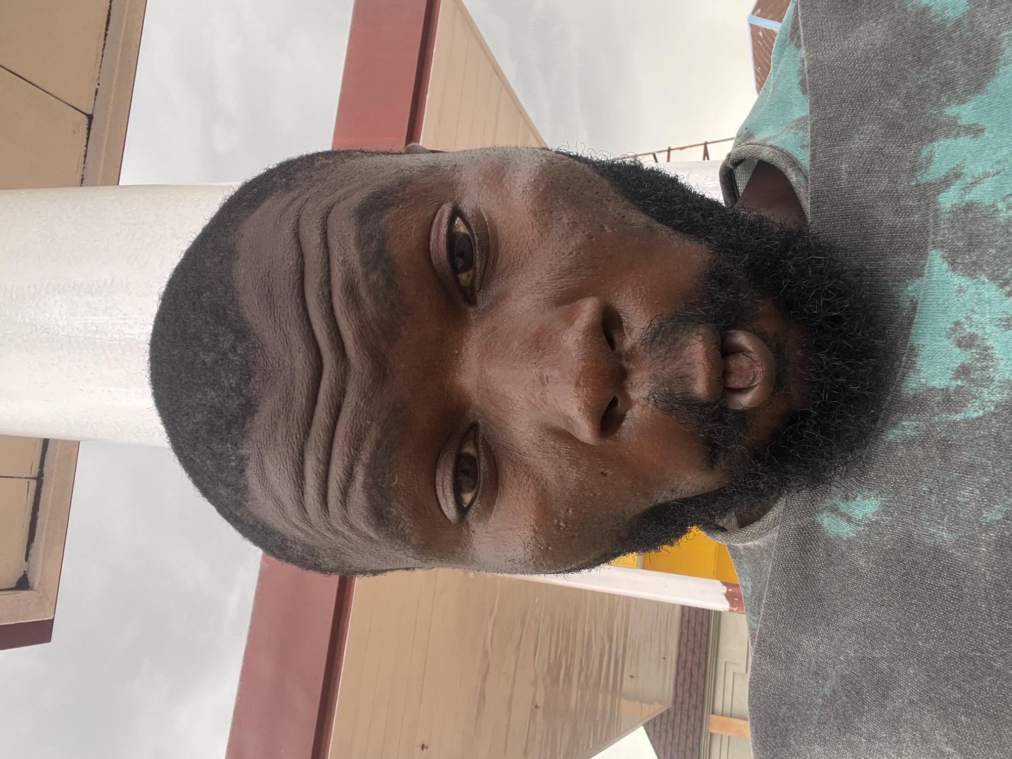 A close-up, portrait-style, slightly angled, outdoor photograph shows a Black man with a shaved head and full beard. The man is wearing a gray shirt with teal splotches. The background consists of beige and red building elements, some white architectural details, and a cloudy sky. It's not possible to determine the exact location of Barakin Lerawi, Nigeria in this image.