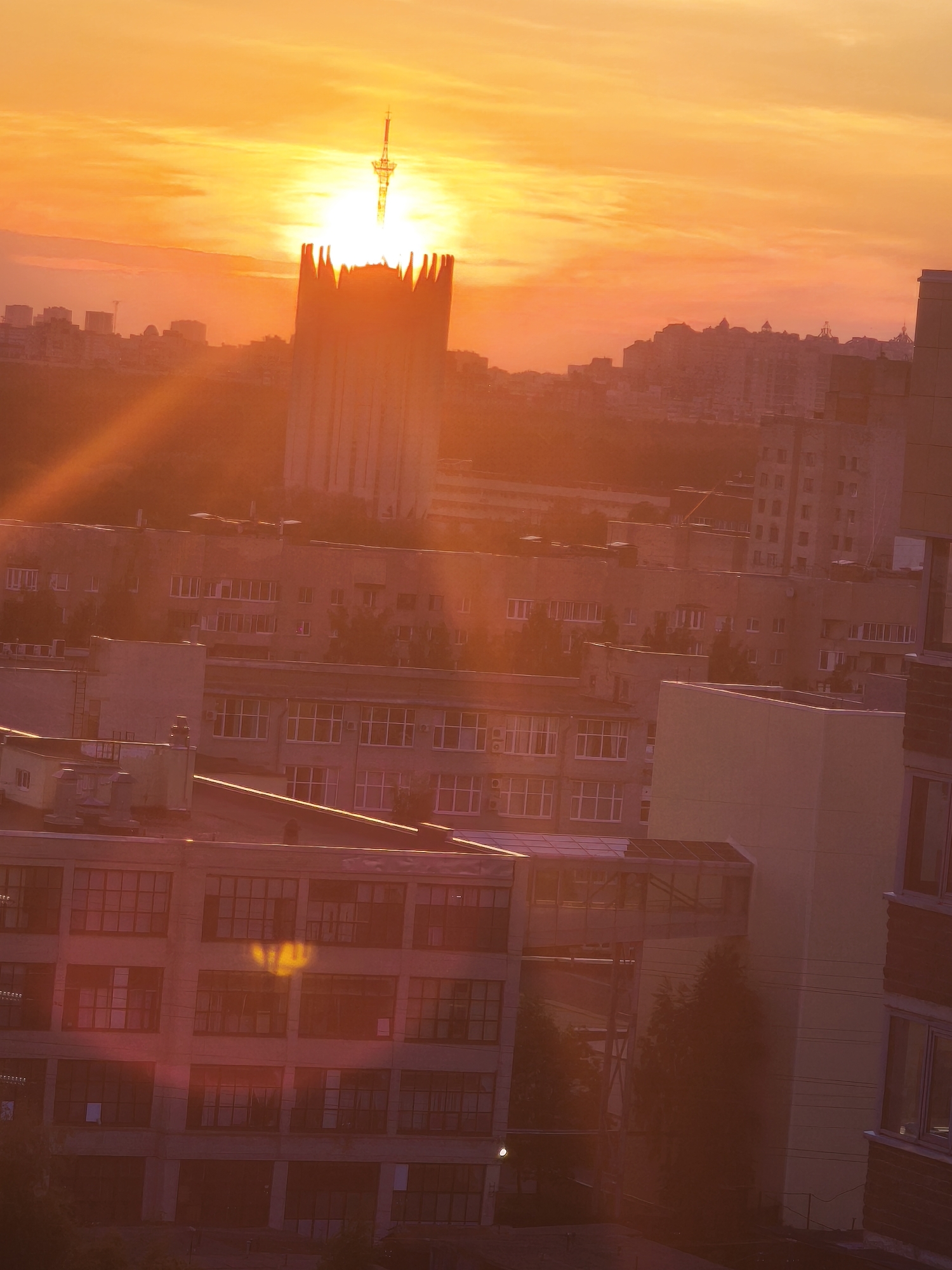 The image shows a cityscape at sunrise or sunset, likely in Saint Petersburg, Russia, based on the presence of the Saint Petersburg TV Tower. The sun is positioned behind the Saint Petersburg TV Tower, creating a silhouette effect. In the foreground and midground, there are multiple buildings, mainly residential apartments, with many windows. The sky is orange and yellow. Light rays from the sun are visible. The image has a hazy, warm tone.