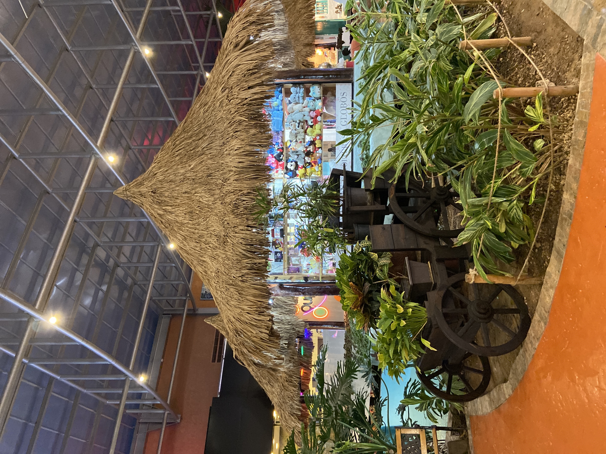 This media file depicts an indoor commercial setting, likely within a shopping mall or large market, featuring a tropical-themed display in Pampatar, Venezuela.
In the foreground, a prominent hut-like structure with a rustic thatched roof, made from dried straw or palm fronds, stands as a central decorative element. Behind this hut, a shop window is visible, brightly lit and filled with a diverse array of colorful stuffed animals and plush toys, suggesting a toy, gift, or novelty store. Partially legible on the shop window is the Spanish word "GLOBOS," meaning "balloons."
To the right of the hut, a planter box holds dense, lush green foliage, contributing to the tropical ambiance. Within this planter, a decorative rustic wooden cart or wagon with large, dark, spoked wheels is positioned among the plants. Some of the plants are supported by wooden stakes tied with string. A distinctive reddish-orange wall is visible on the far right edge of the frame.
The background features a high, architectural ceiling structure composed of metallic beams and transparent or translucent panels, possibly a skylight or an atrium roof. Several small, bright lights are embedded within these beams, indicating an indoor environment or evening time. The scene is devoid of people, focusing instead on the themed decor and commercial offerings.