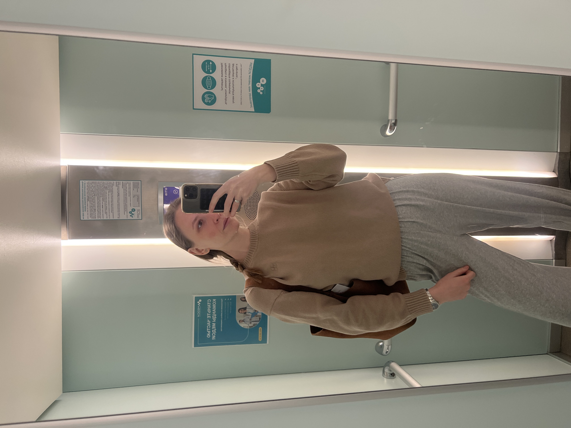 A woman is taking a selfie in what appears to be a public restroom. She is wearing a light brown sweater and gray sweatpants, holding her phone up to capture her reflection in the mirror. The restroom has a modern, minimalist design with light blue walls and a white countertop. There are signs on the wall, one with medical-related information and another with COVID-19 safety guidelines. The text on one sign is partially written in Russian, specifically mentioning the city of Khimki. There is a silver grab bar visible near the countertop. The restroom appears clean and well-maintained.
