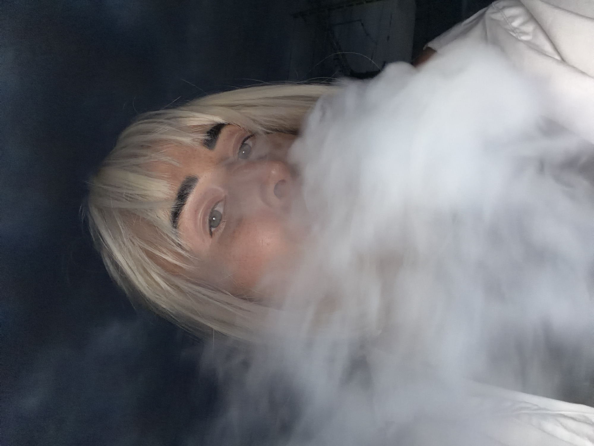 In the image, a person with blonde hair and bangs is partially obscured by a cloud of white vapor, possibly smoke. The person's eyes are visible, and they appear to be looking forward. The background is dark and somewhat blurry, with hints of what might be structures or industrial elements, suggesting an urban or industrial setting, potentially near power lines or scaffolding. The lighting is dim, casting a shadowy ambiance. The image does not contain any explicit references to the city of Sochi, Russia, or any other specific location.