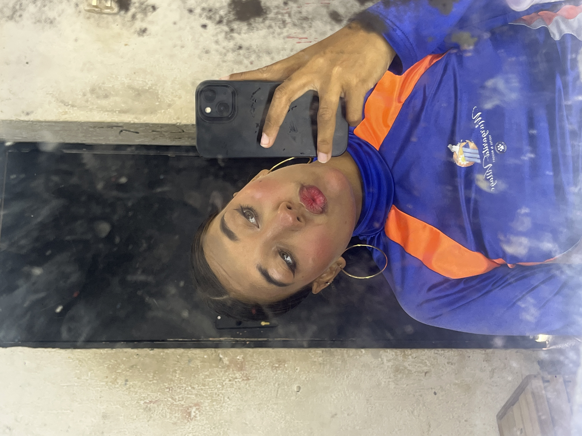 A person is taking a selfie in a mirror. The person is wearing a blue and orange athletic shirt. They are holding a black smartphone. The mirror and surrounding area appear dirty. There is no indication of this picture's location.