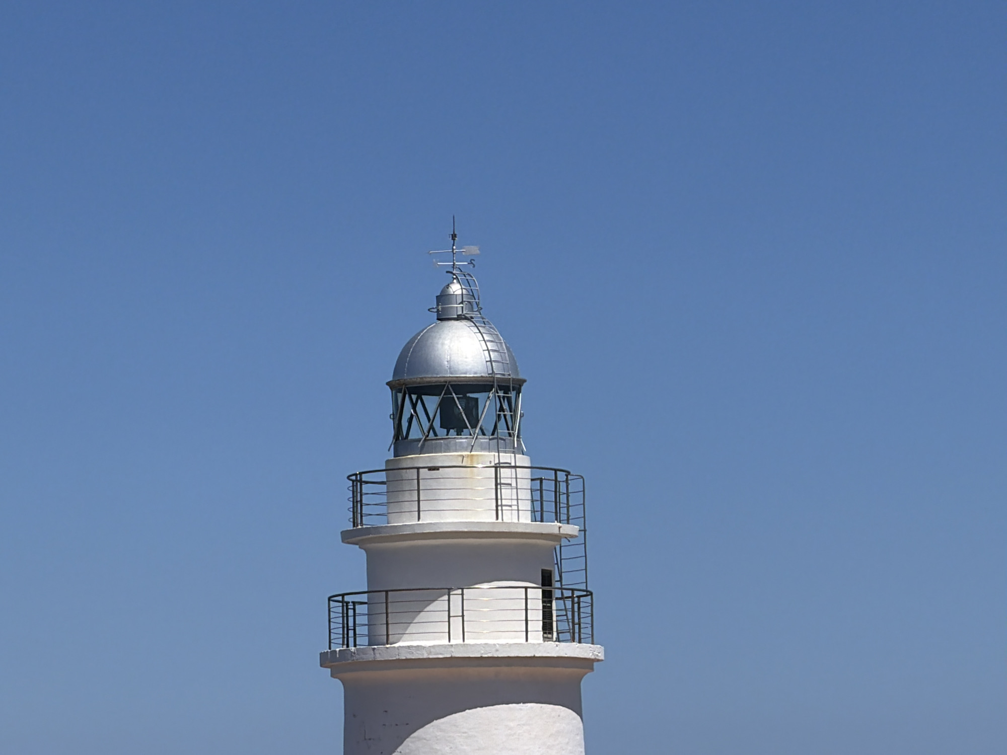 A medium shot depicts the upper portion of the Capdepera Lighthouse in Capdepera, Spain, under a clear blue sky. The lighthouse is white with a silver-colored dome at the top. It features two viewing balconies with black metal railings. A metal weather vane sits atop the dome. A small window is visible on the main section of the lighthouse below the upper balcony.
