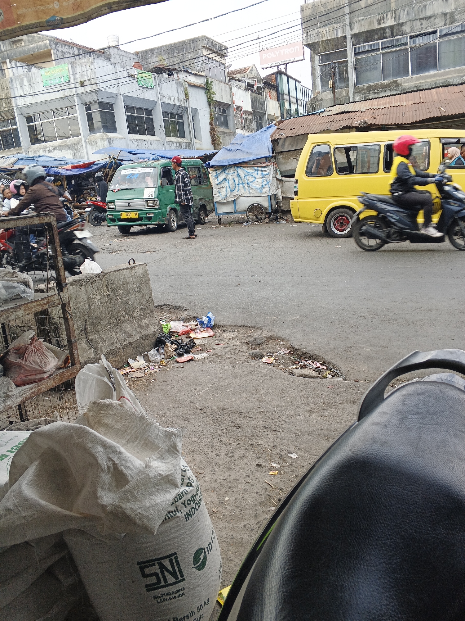 A street scene in Sukabumi, Indonesia, shows a busy market area with vehicles and pedestrians. A green minivan with license plate "F 1951 ST" is parked on the right side of the road, facing left. A man in a checkered shirt and red hat stands next to it, facing the van. To the right of the minivan, a yellow bus is visible, with people inside. A motorcycle is in motion on the right, with a rider wearing a red helmet and a yellow jacket. In the foreground, a motorcycle seat is partially visible. The ground is asphalt, with some debris and trash scattered around. To the left of the minivan, a rack with bags of white material is present. Buildings line the background, appearing to be shops and commercial establishments. The sky is overcast.
