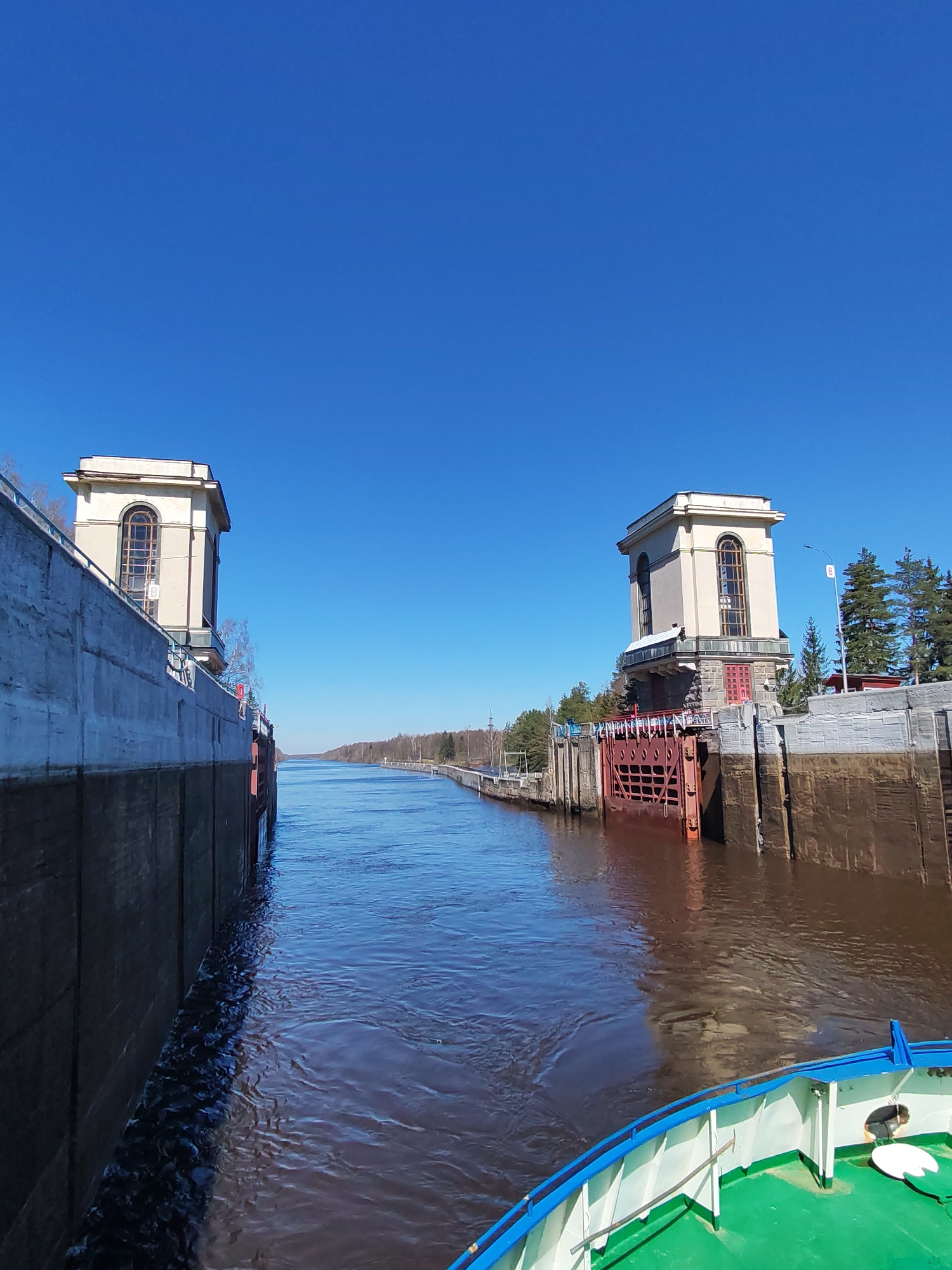 The image shows a waterway, possibly a canal or river, passing through a lock system. The lock is flanked by two structures: towers with arched windows, possibly control towers for operating the lock gates. The gates themselves are red and appear to be closed. The water in the waterway is a murky brown color. A section of a boat's deck is visible in the foreground, suggesting the photo was taken from a boat moving through the lock. The sky is clear blue, indicating a sunny day. Trees line the banks of the waterway in the distance. The location is Unknown, Unknown.