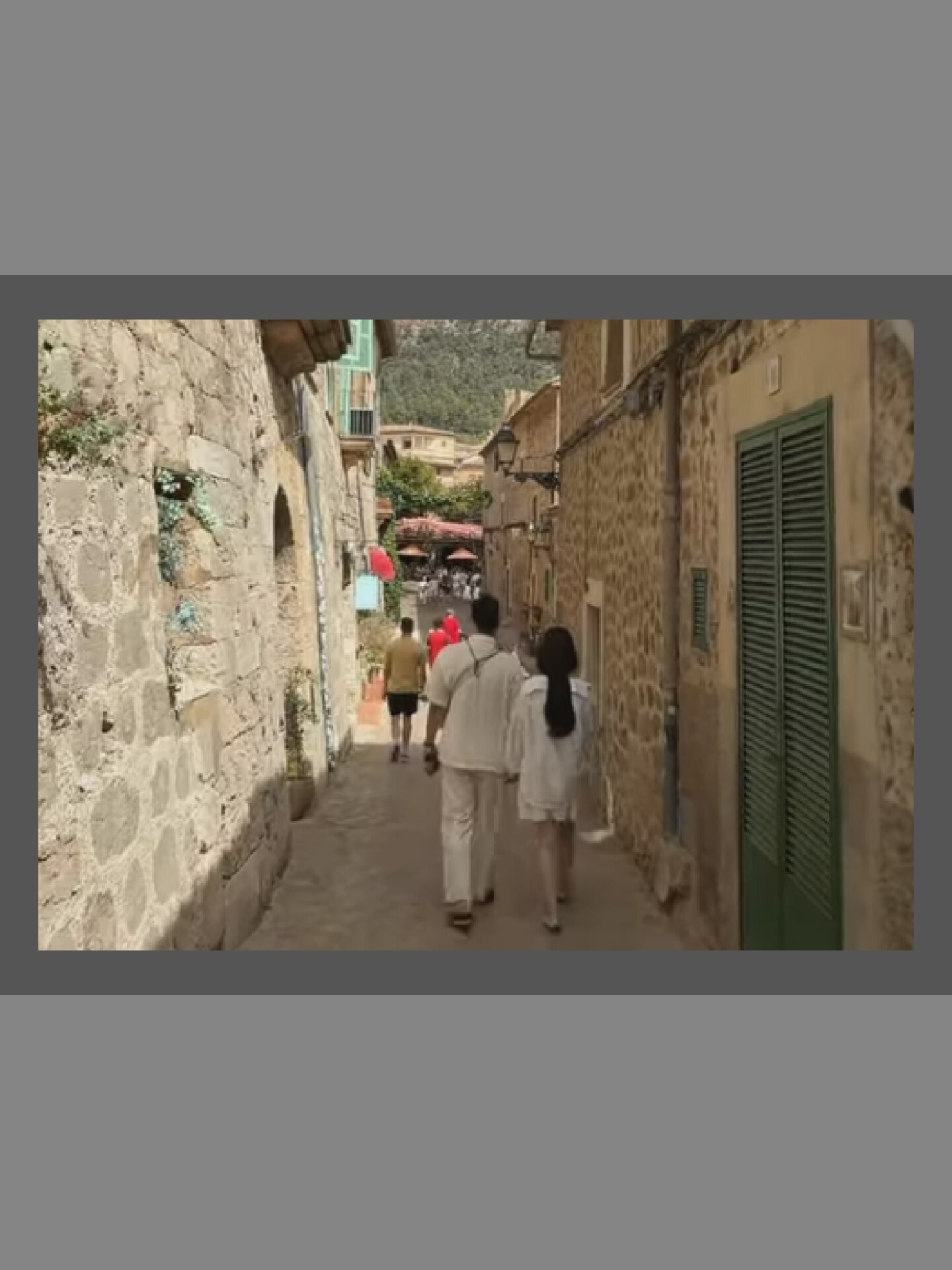 A man and a woman are walking down a narrow, cobblestone street. The man is wearing a light-colored shirt and pants, and the woman is wearing a white dress. They are walking away from the camera, towards the distance. In the background, there are stone buildings with green shutters and some foliage. Further down the street, there are other people walking and sitting at tables outside. The scene appears to be a historic European town.