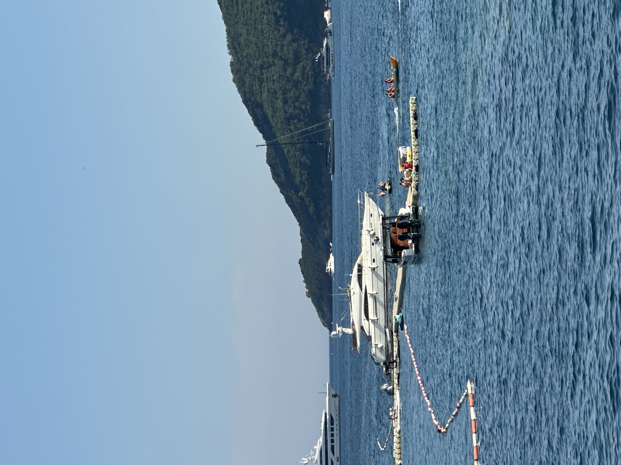 The image depicts a sunny day at a coastal location, likely Budva, Montenegro, characterized by a clear blue sky and calm blue waters. The scene is dominated by several boats, including a large white yacht moored at a dock and another smaller white boat visible in the background.
The dock is bustling with activity. People are seen relaxing on floating inflatable devices and sitting on the dock itself. One inflatable, resembling a banana boat, has several individuals riding on it. Another smaller inflatable raft is also present with people on it. The activity suggests recreational water sports or leisure time.
In the background, a lush green, tree-covered hill slopes down towards the water, providing a scenic backdrop. The time of day appears to be daytime, with bright sunlight casting minimal shadows, indicating a warm and clear day. The overall mood is relaxed and leisurely, typical of a summer vacation or a day spent enjoying water activities at a seaside resort. No visible text is present in the image.