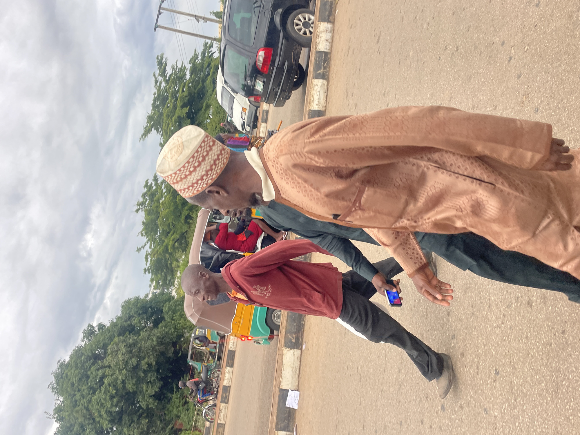 In Barakin Lerawi, Nigeria, under an overcast sky, three men are captured in a mid-stride procession across a paved road. The man in the foreground, clad in a light brown, intricately patterned kaftan and a white and red embroidered cap, walks towards the right of the frame, his left hand visible. Adjacent to him and slightly behind, another man in a dark garment walks with his right hand holding a smartphone. Further back, a third man in a maroon hooded sweatshirt and dark trousers, with a shaved head, is also in motion, facing towards the left. A yellow and green auto-rickshaw is visible to the left, its open doors revealing passengers. A red motorcycle with a rider is also in the background, along with a black SUV and a white van. The road is bordered by a curb with alternating black and white markings, and a line of lush green trees forms the backdrop. The scene suggests a typical streetscape in Nigeria, with vehicular traffic and pedestrians navigating the thoroughfare.