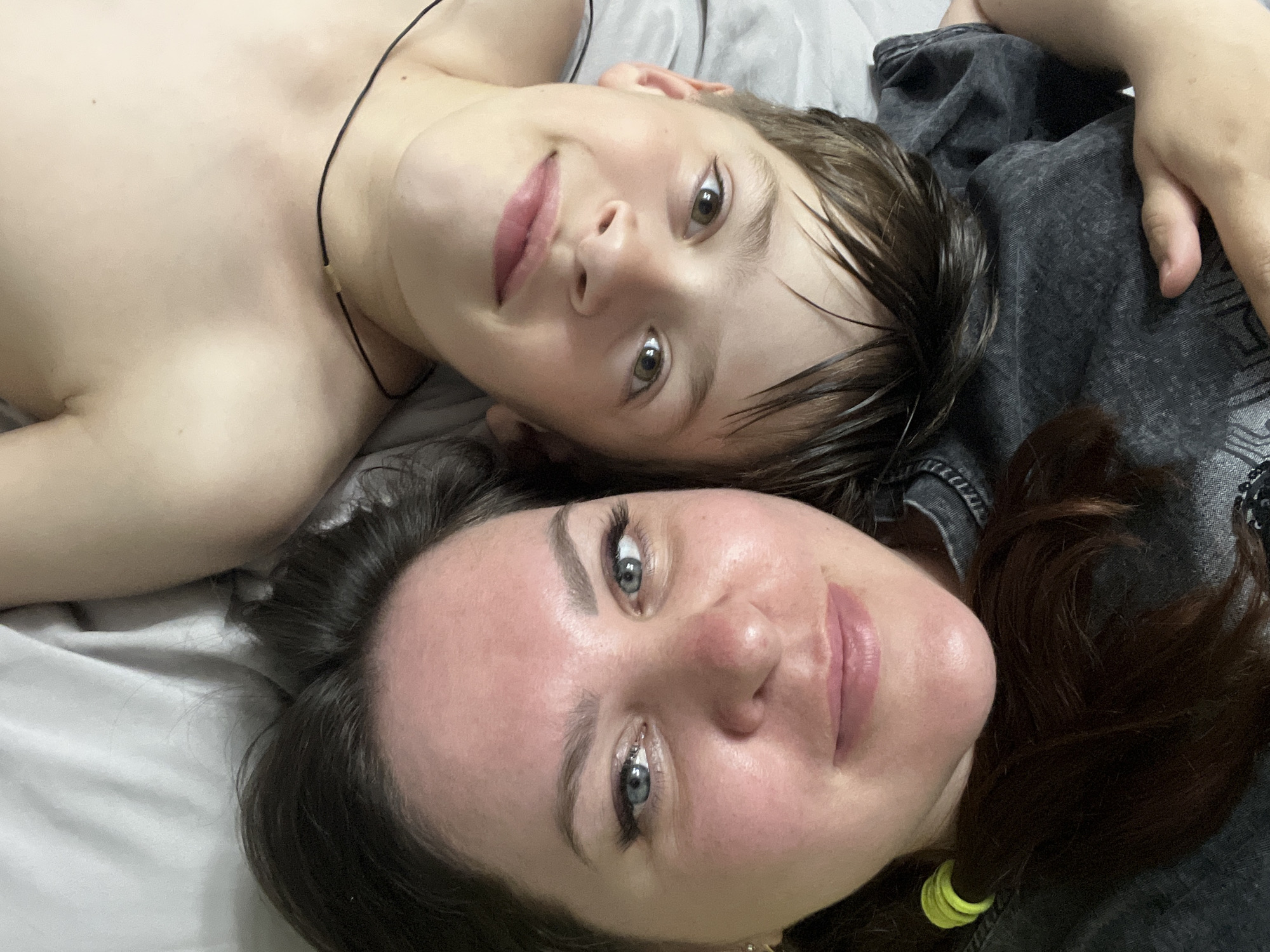A woman and a boy lie on a gray surface, likely a bed, looking at the camera. The boy is on the left side of the image and has light brown hair. The woman is on the bottom of the image, and has dark hair and blue eyes. She is wearing a dark gray top. The background is a plain gray color. There are no indications of the location being in the city of Noviy, Russia.