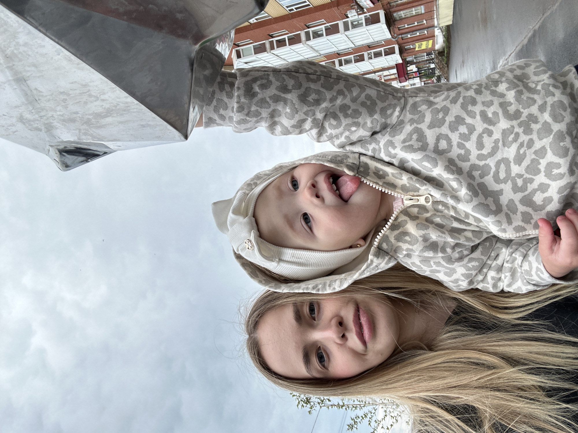 The image, taken from a low-angle perspective, features an adult woman and a baby outdoors in Азинский, Russia. The woman, positioned closer to the camera, has long blonde hair, blue eyes, and appears to have braces on her teeth; she looks directly forward with a calm expression. Directly above her, a baby, wearing a grey and white leopard print hooded jacket and a white beanie, playfully sticks out its tongue, looking up towards the camera with blue eyes.
A large, angular metallic structure occupies the upper left portion of the frame, reflecting light. In the background, under a cloudy sky, are multi-story residential buildings with multiple windows and balconies, typical of an urban or suburban environment. The ground behind the baby appears wet, suggesting recent rain or damp conditions. On one of the distant buildings, a small, yellow sign with unidentifiable text is barely visible. The overall impression is a casual, intimate moment captured on a cloudy day.