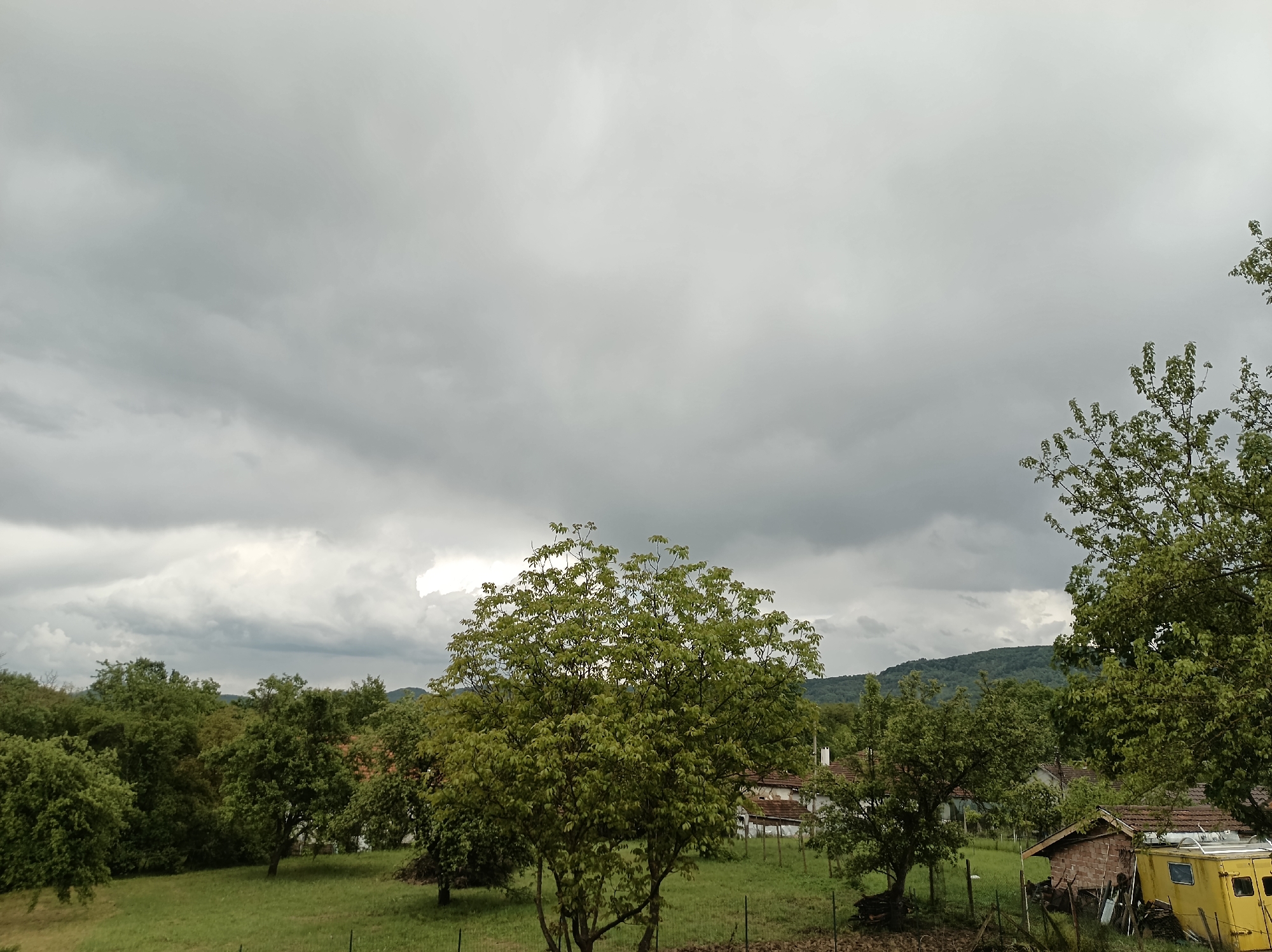The image shows a rural scene, potentially in or near Mihaylovtsi, Bulgaria. The sky is overcast with heavy, gray clouds suggesting an imminent storm. The foreground is dominated by a green field with several trees scattered throughout. One large, bushy tree stands prominently in the center. In the midground, glimpses of buildings with tiled roofs can be seen partially obscured by trees. To the right, a small, yellow trailer is parked near a brick structure with a tile roof. A fence runs along the bottom portion of the image, partially hidden by vegetation. In the background, a slightly elevated, tree-covered terrain is visible. The scene appears to be a residential area blended with a natural landscape on a cloudy day. There is no visible presence of people, no specific actions.
