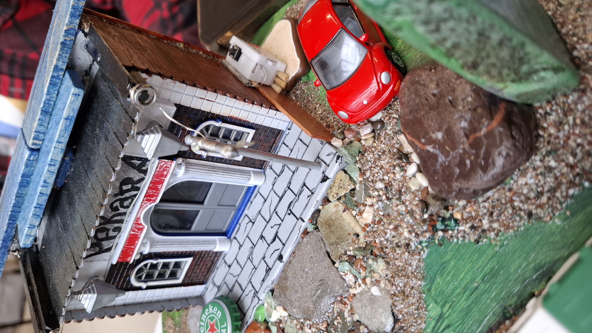 A diorama presents a miniature, meticulously detailed scene. It features a model building that mimics a European architectural style, possibly inspired by structures in Budva, Montenegro. The building has a brick facade and tiled roof, with window frames painted white and red. The rooftop is adorned with a small, white electric box. Adjacent to the building is a gravel-covered ground, speckled with small rocks and miniature items, including a red toy car, green bottle caps with a Heineken logo. A large, smooth rock is also visible, and the artificial turf suggests a grassy area. The diorama is positioned on a larger base, giving the impression of a landscaped environment.