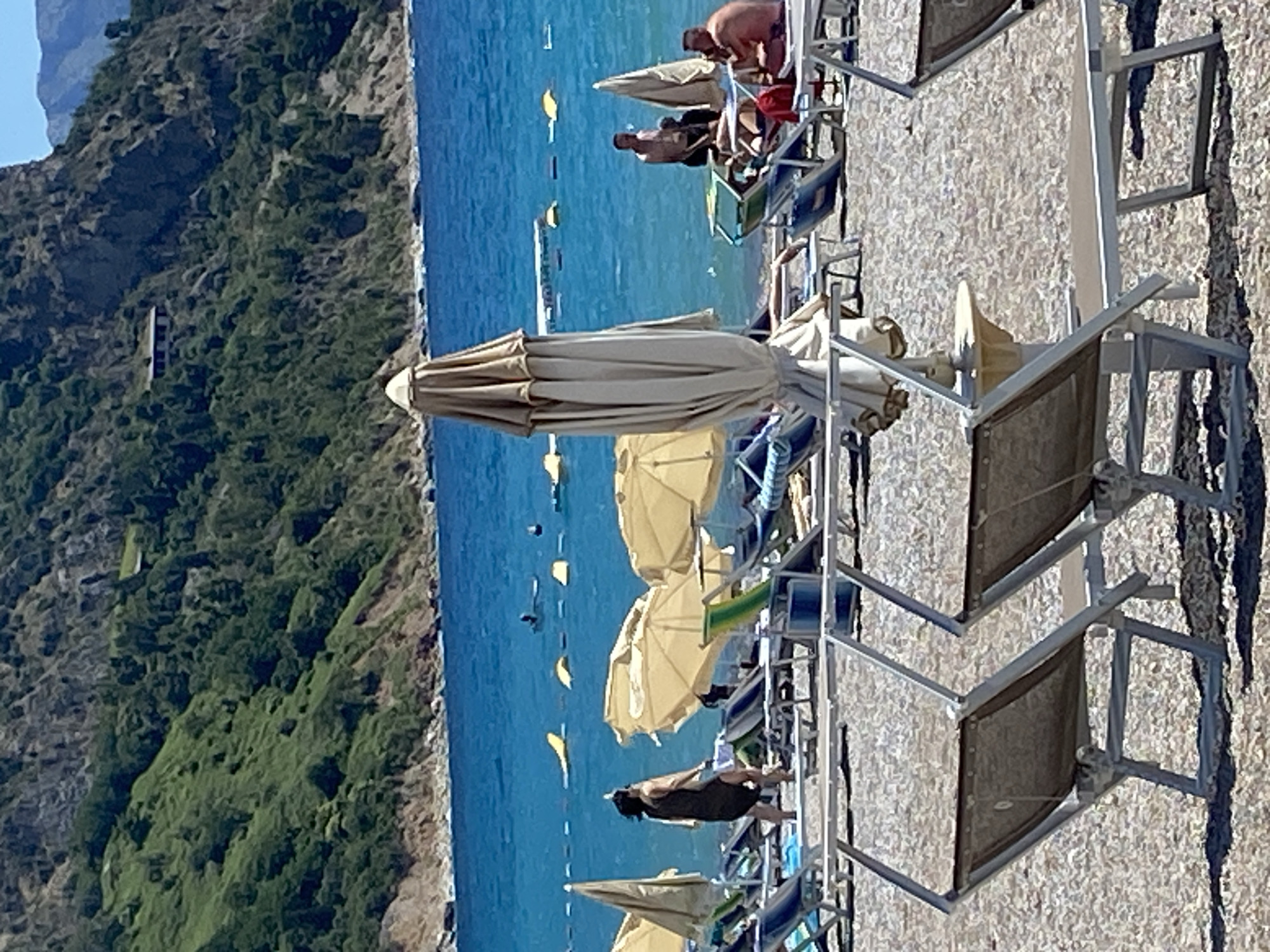 The image depicts a sunny day at a beach in Prijevor, Montenegro. The foreground is dominated by rows of empty, light-colored sun loungers and closed beach umbrellas on a pebble-covered shore. The sea is a vibrant blue, with small waves lapping at the shore. In the mid-ground, several people are visible. One woman in a dark dress walks towards the sea, while a group of men are seated on loungers near the water's edge. Further out in the sea, some inflatable buoys are scattered. The background features a steep, lush green hillside dotted with trees and a few buildings. The sky is clear and blue, indicating fair weather.
