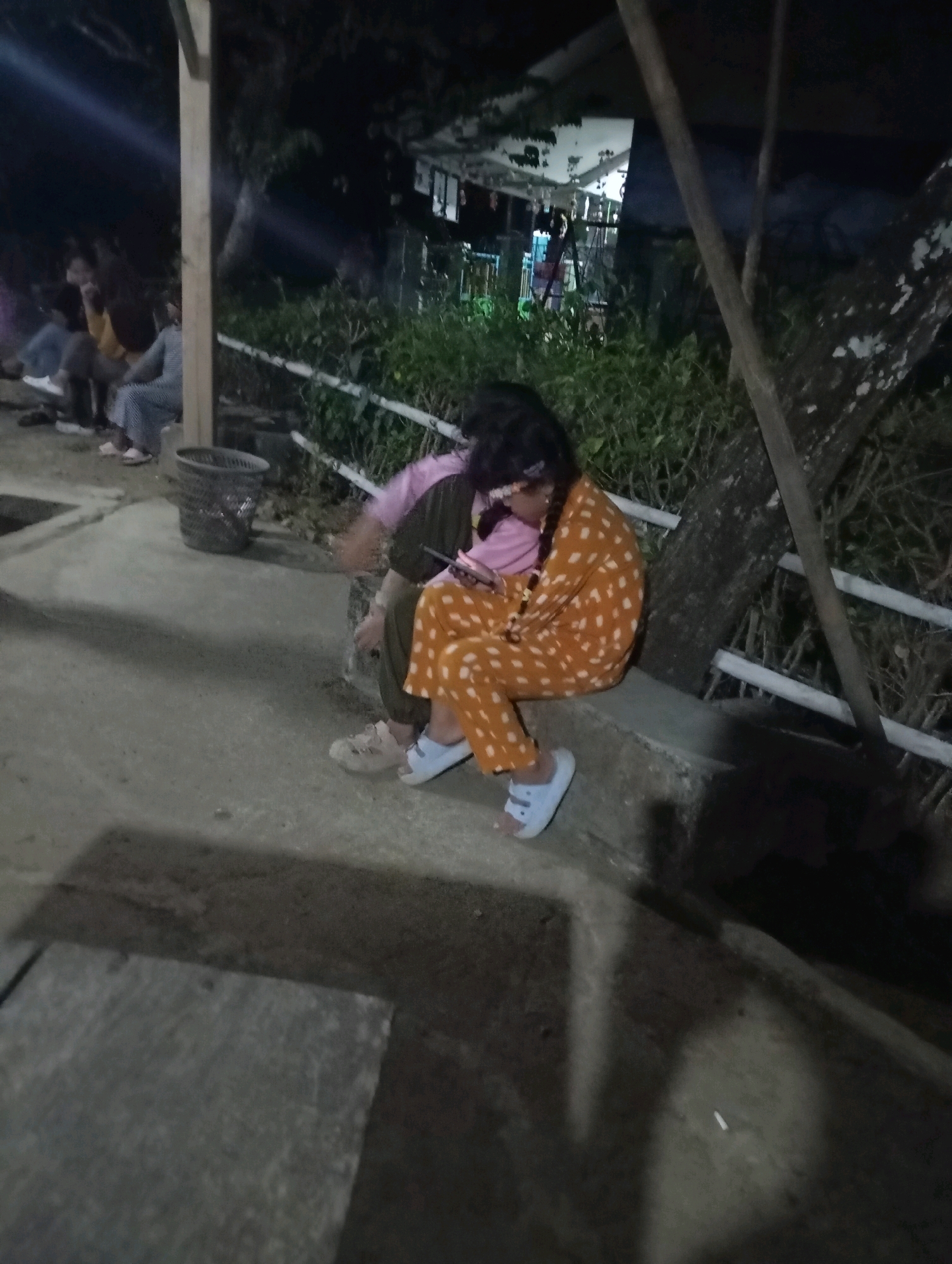 The image depicts a nighttime scene in what appears to be an outdoor setting, potentially a park or a communal area, with hints of Indonesian urban elements such as a trash can that resembles those found in Indonesian public spaces. In the foreground, a person is seated on a low concrete barrier, their legs bent and feet resting on the ground. They are wearing a brightly colored orange jumpsuit with white polka dots and white slide-on sandals. This individual is holding a smartphone and appears to be looking at it. Behind this person, another individual is partially visible, wearing a pink top and darker clothing. In the background, several other people are seated or standing, their figures obscured by the low light and distance. They are gathered near a wooden structure and a fence with white railings. Lush green foliage is visible behind the fence, and a large tree trunk is prominent on the right side of the image. The ground is concrete, with some shadows cast from the lighting, suggesting artificial illumination from overhead or nearby sources. The overall atmosphere is casual and relaxed, with individuals gathered outdoors in the evening. The presence of the trash can and the style of the railing are consistent with elements found in public spaces in Ketepung, Indonesia.