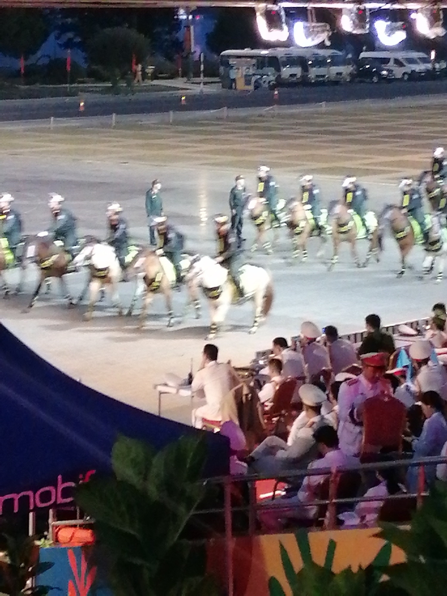 The image depicts a large outdoor event, likely a parade or ceremony, taking place in an open paved area. In the foreground, a blue tarp with pink lettering, partially obscured by greenery, is visible. Behind this, a group of spectators are seated, some wearing uniforms and hats. The main focus of the scene is a procession of horse riders, dressed in dark uniforms and helmets, on horseback. The horses appear to be moving. In the background, several white and dark-colored buses are parked. The overall atmosphere suggests a public gathering, possibly at a stadium or large square in Da Lat, Vietnam.